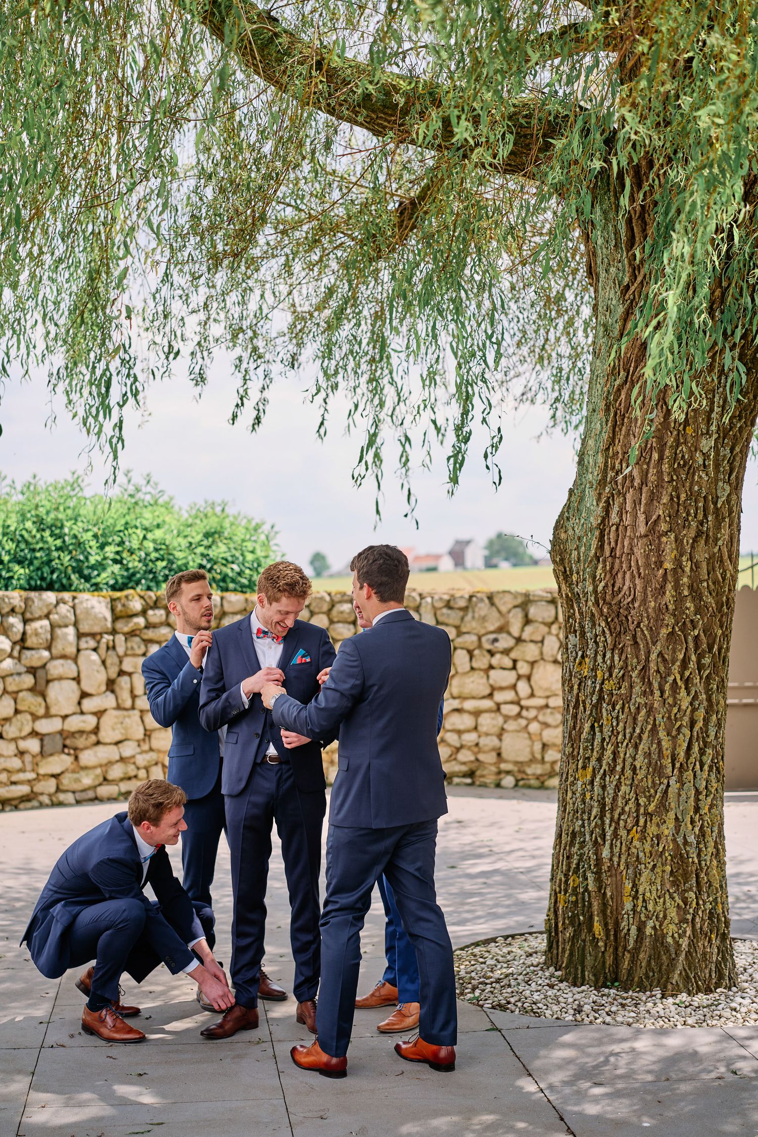 Groomsmen in navy suits gather playfully under a weeping willow tree during a wedding photo session.