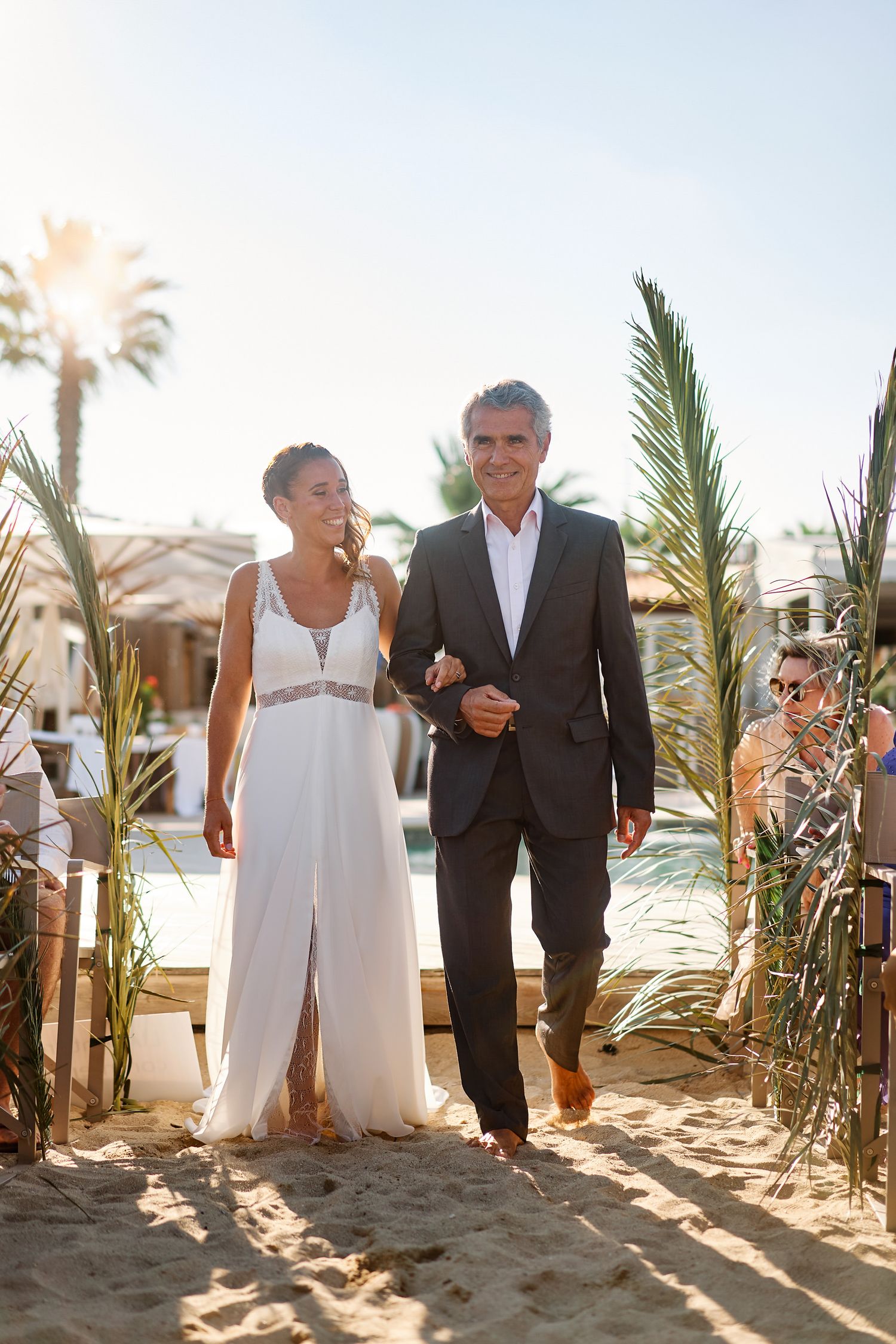 A couple walks together on a sandy beach with palm trees and resort buildings in the background during sunset.