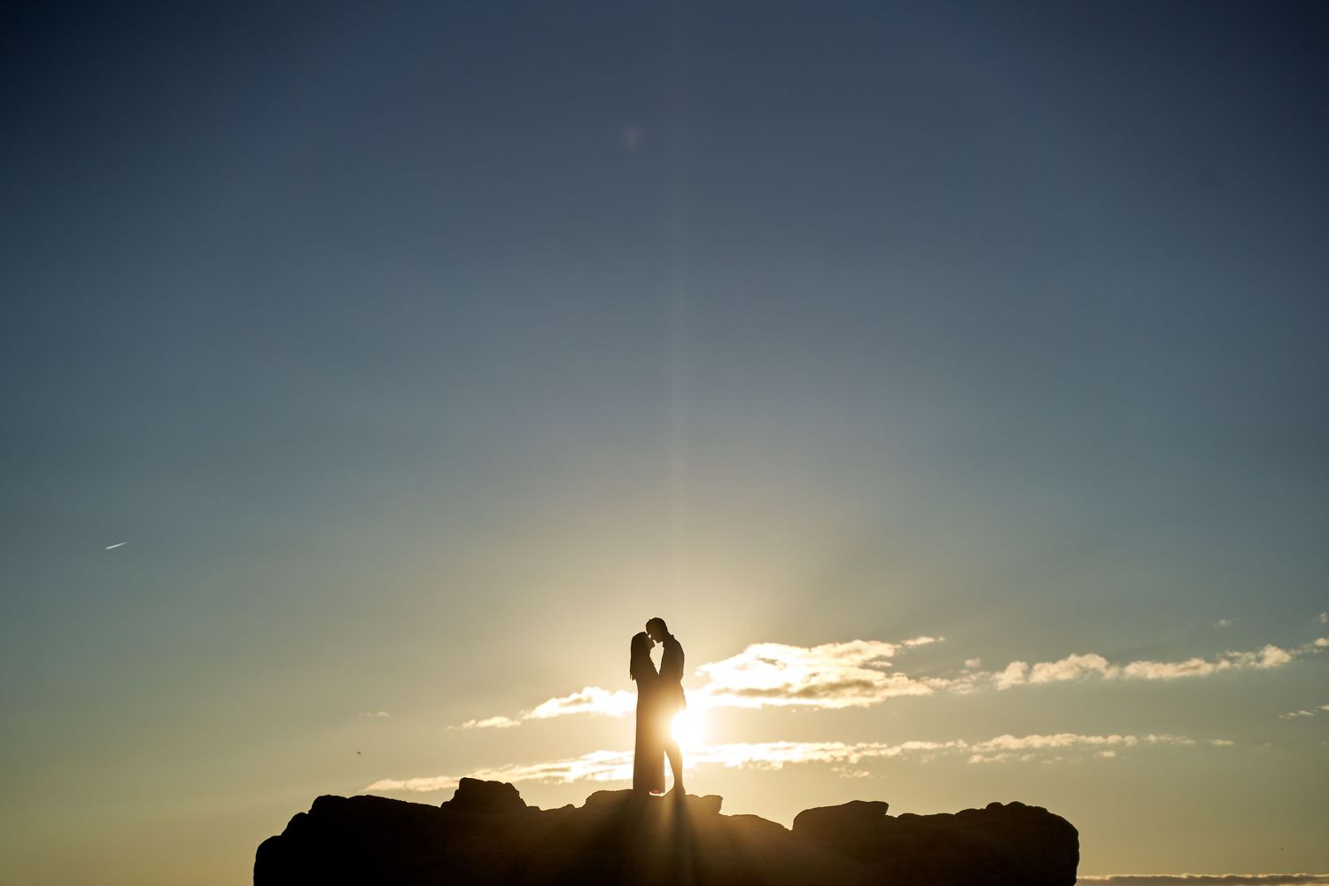 A silhouetted figure stands on rocky cliffs against a dramatic sunset sky with rays of light bursting through clouds.