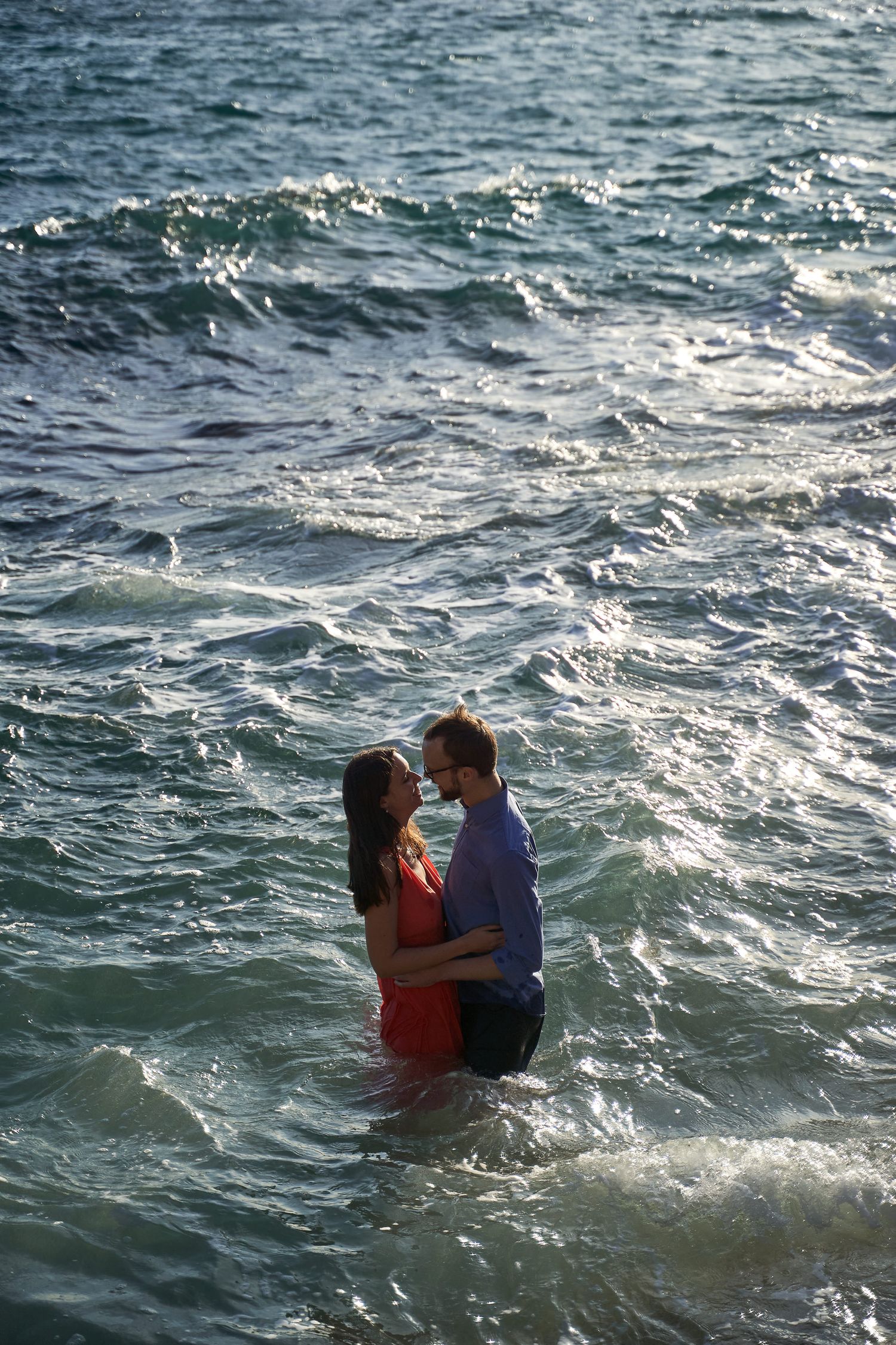 Couple embracing in ocean waves during sunset photoshoot.
