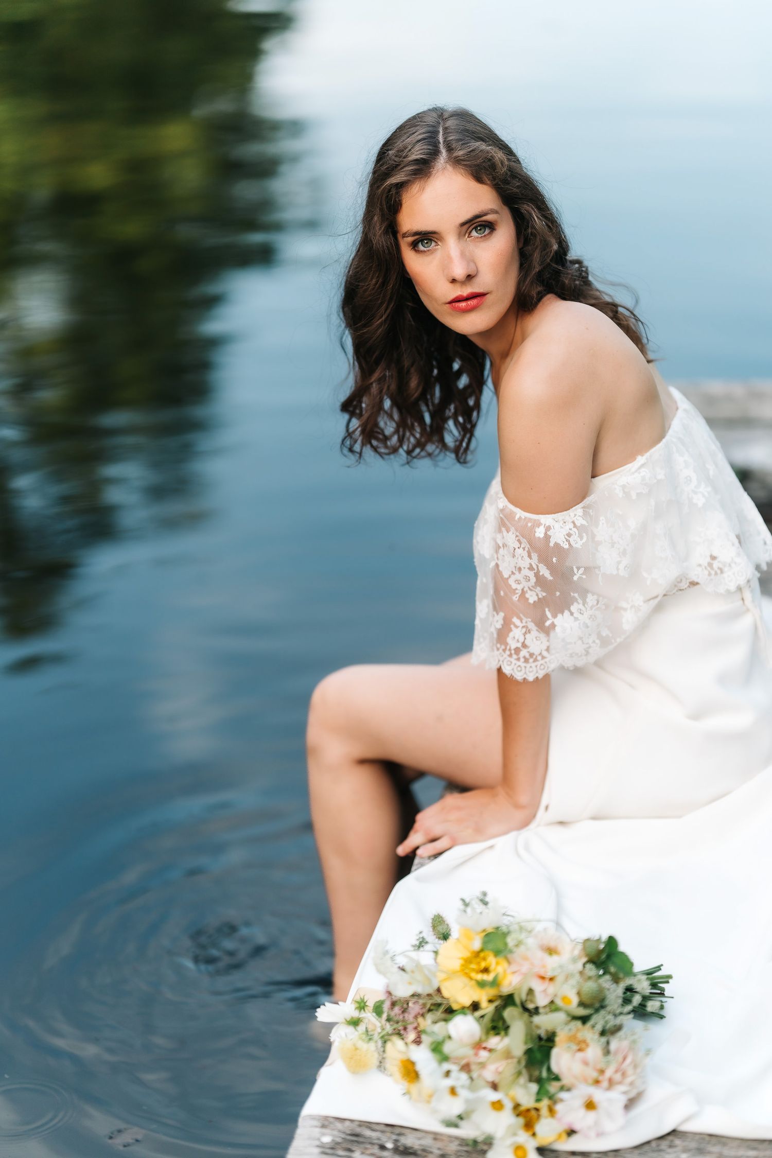 A romantic bridal portrait series by the water featuring a white lace dress and yellow floral bouquet.