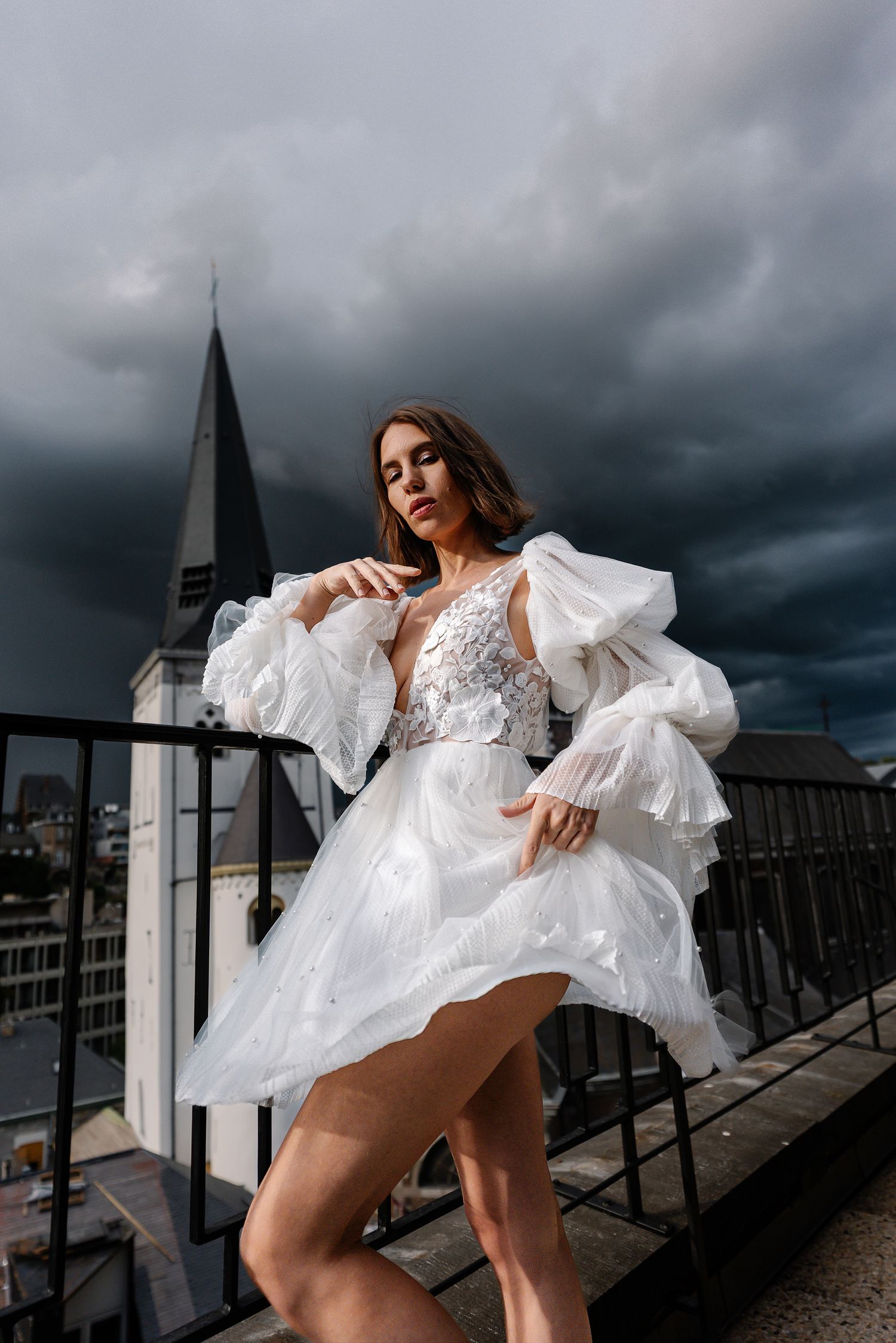 A figure in a flowing white dress poses dramatically against a stormy sky on an urban rooftop.
