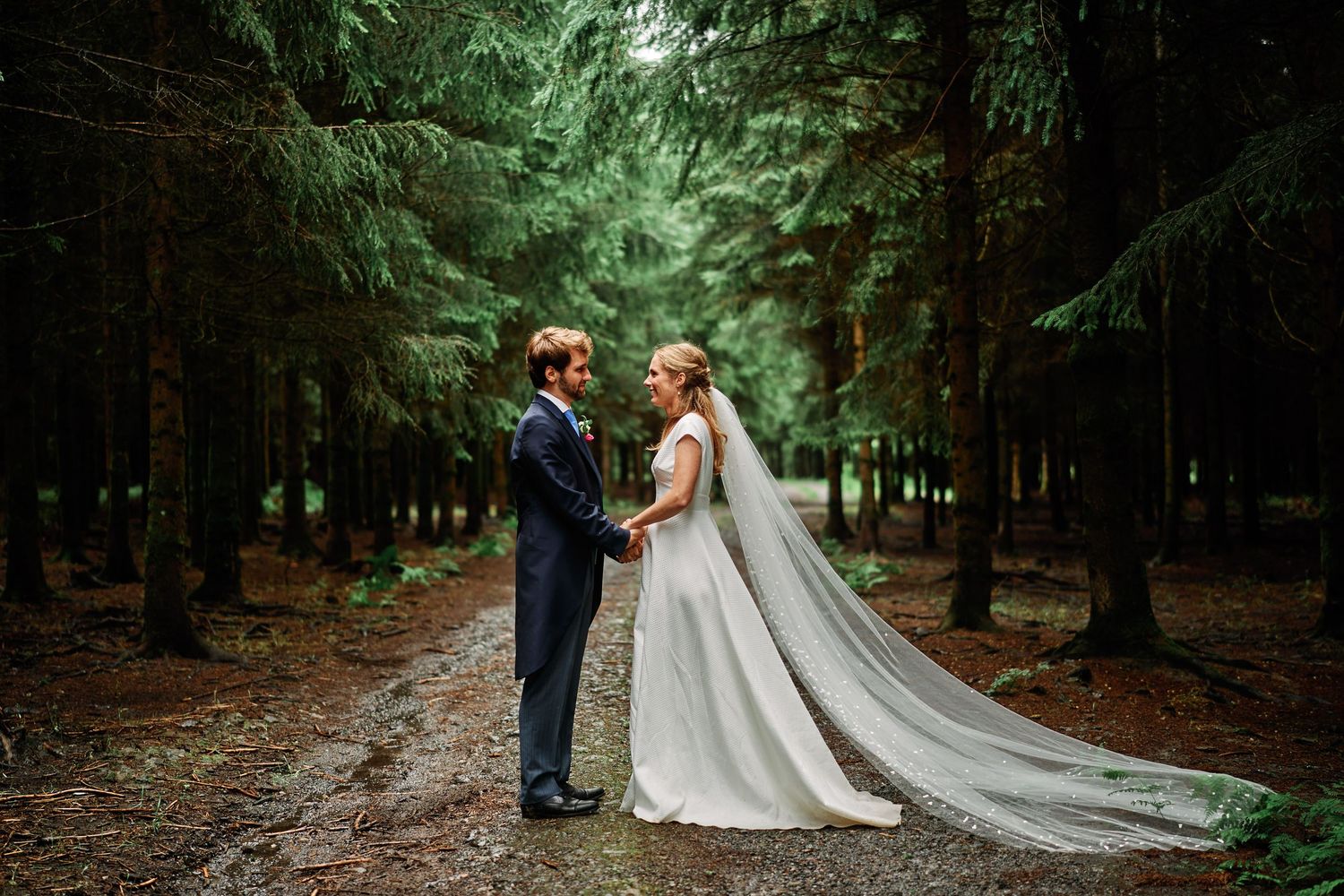 A couple in wedding attire shares intimate moments along a tree-lined forest path during their outdoor ceremony.