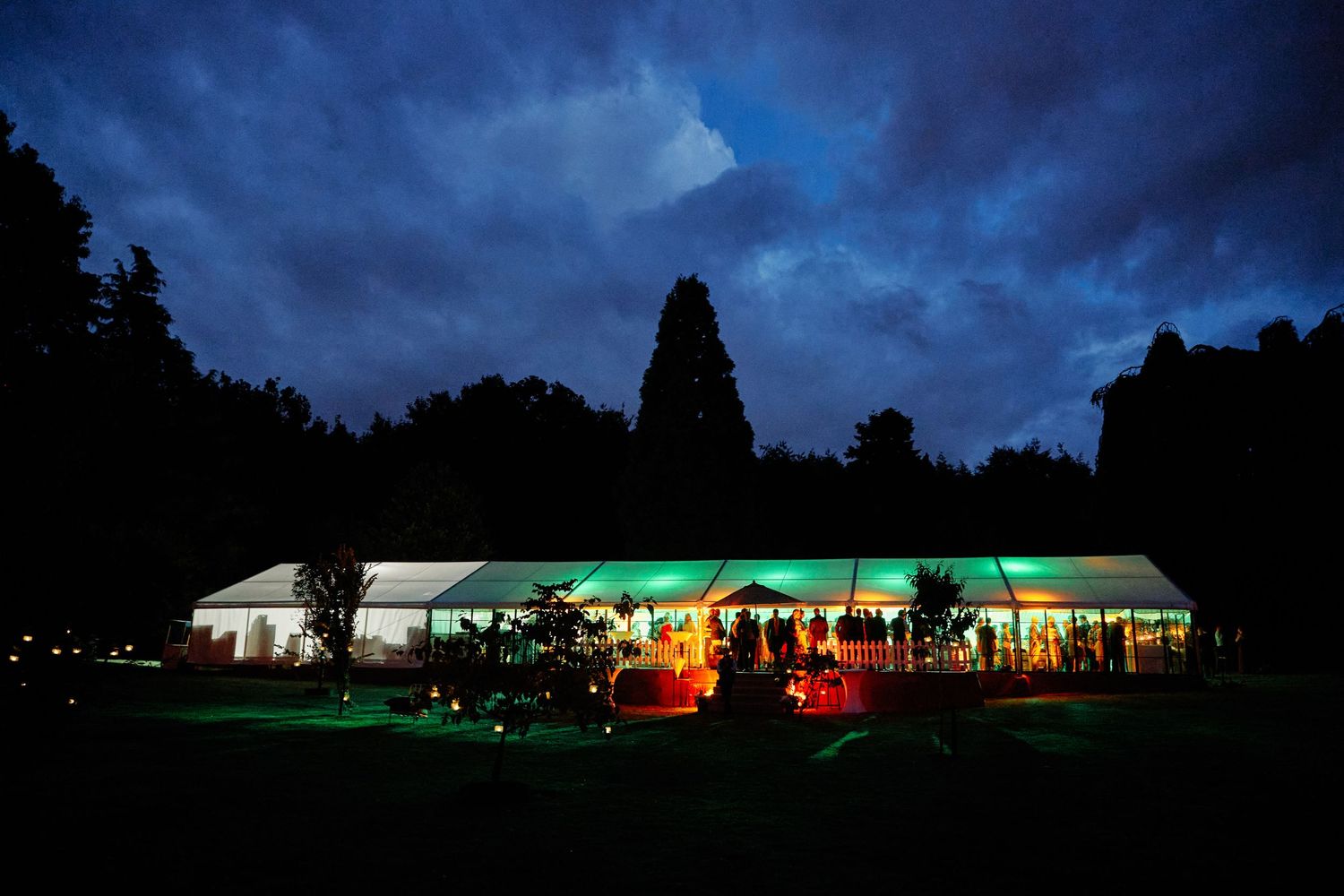 Illuminated tent glows green against dark stormy sky with silhouetted trees in the background.