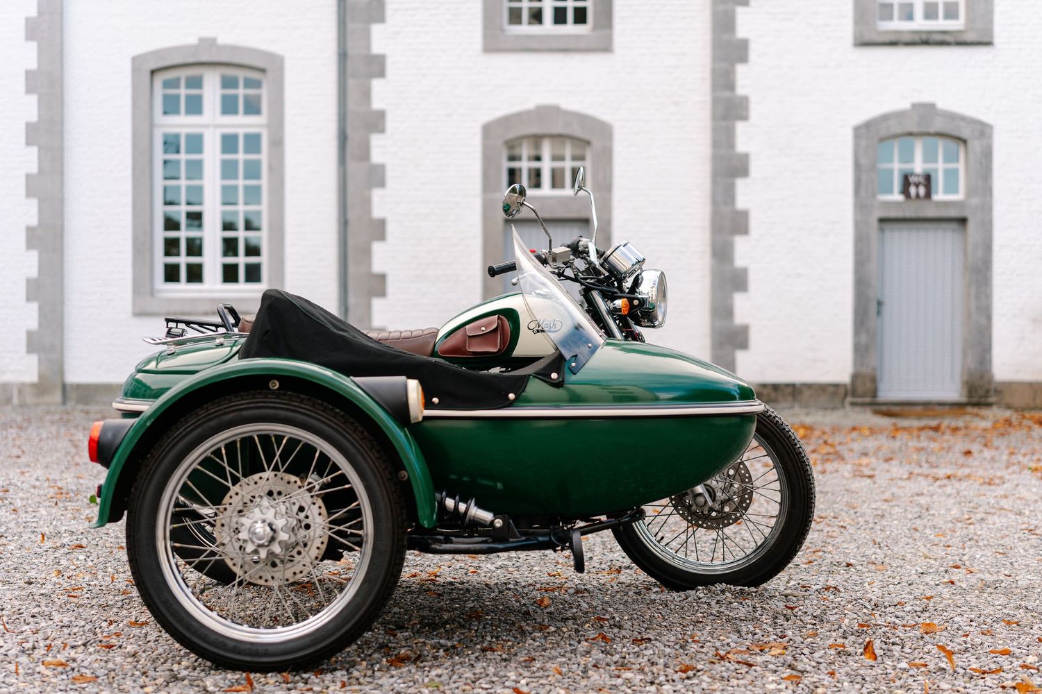 A vintage green motorcycle with sidecar parked in front of a classic white stone building with arched windows.