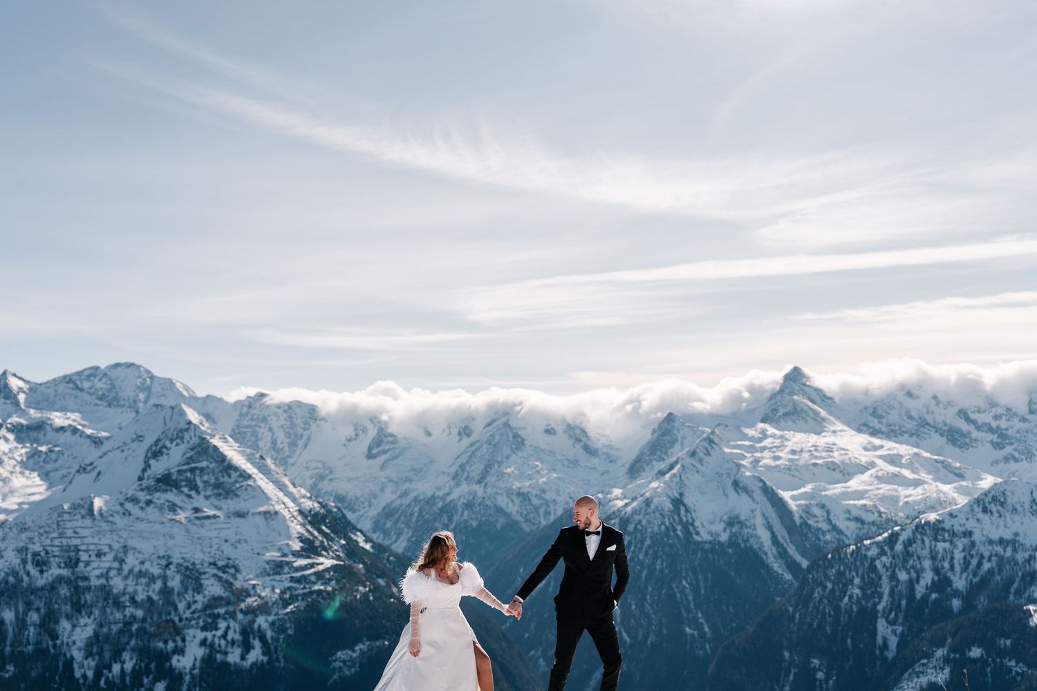 A couple in wedding attire runs together on a snowy mountain peak with dramatic snow-capped Alps in the background.