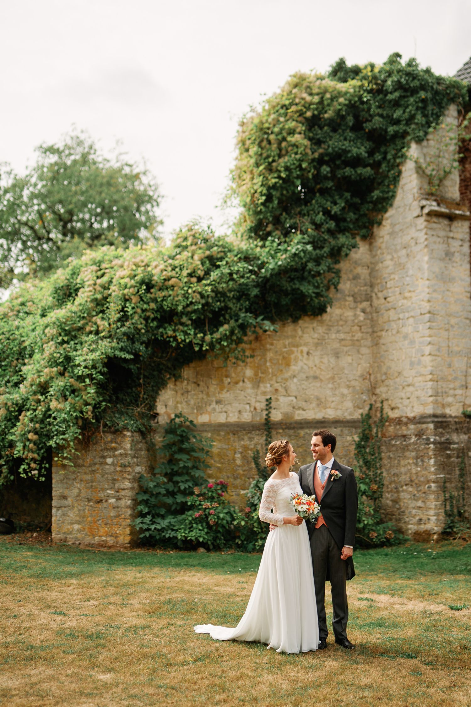 A couple shares an intimate moment against a historic stone wall surrounded by lush greenery in a romantic outdoor setting.