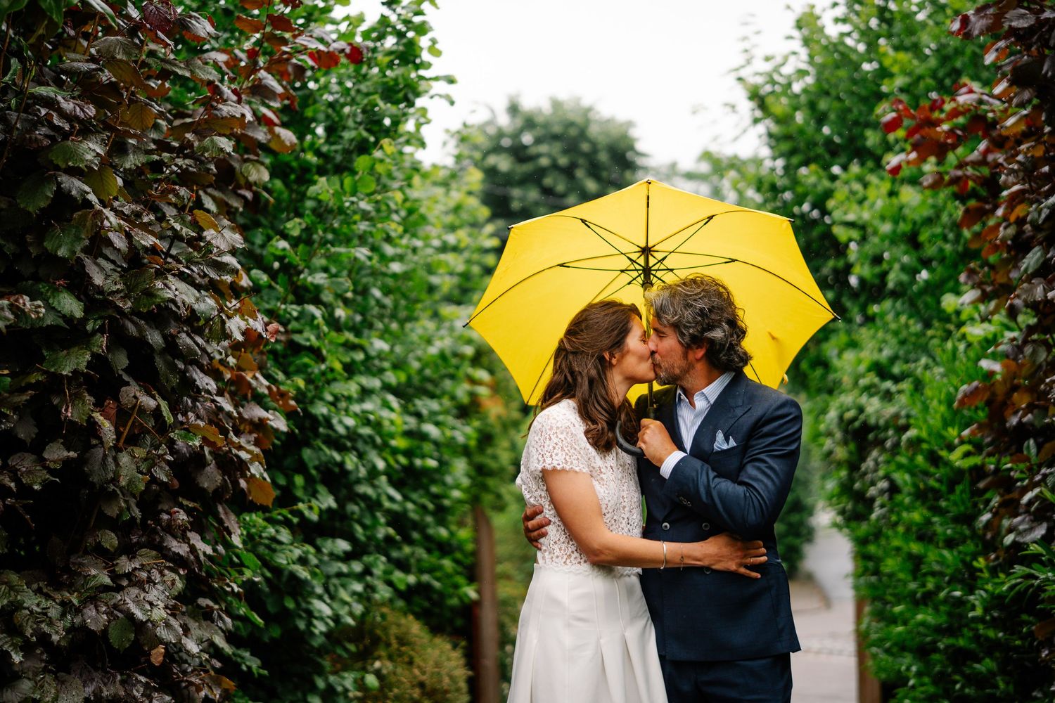A couple shares a romantic kiss under a yellow umbrella in a lush garden setting.