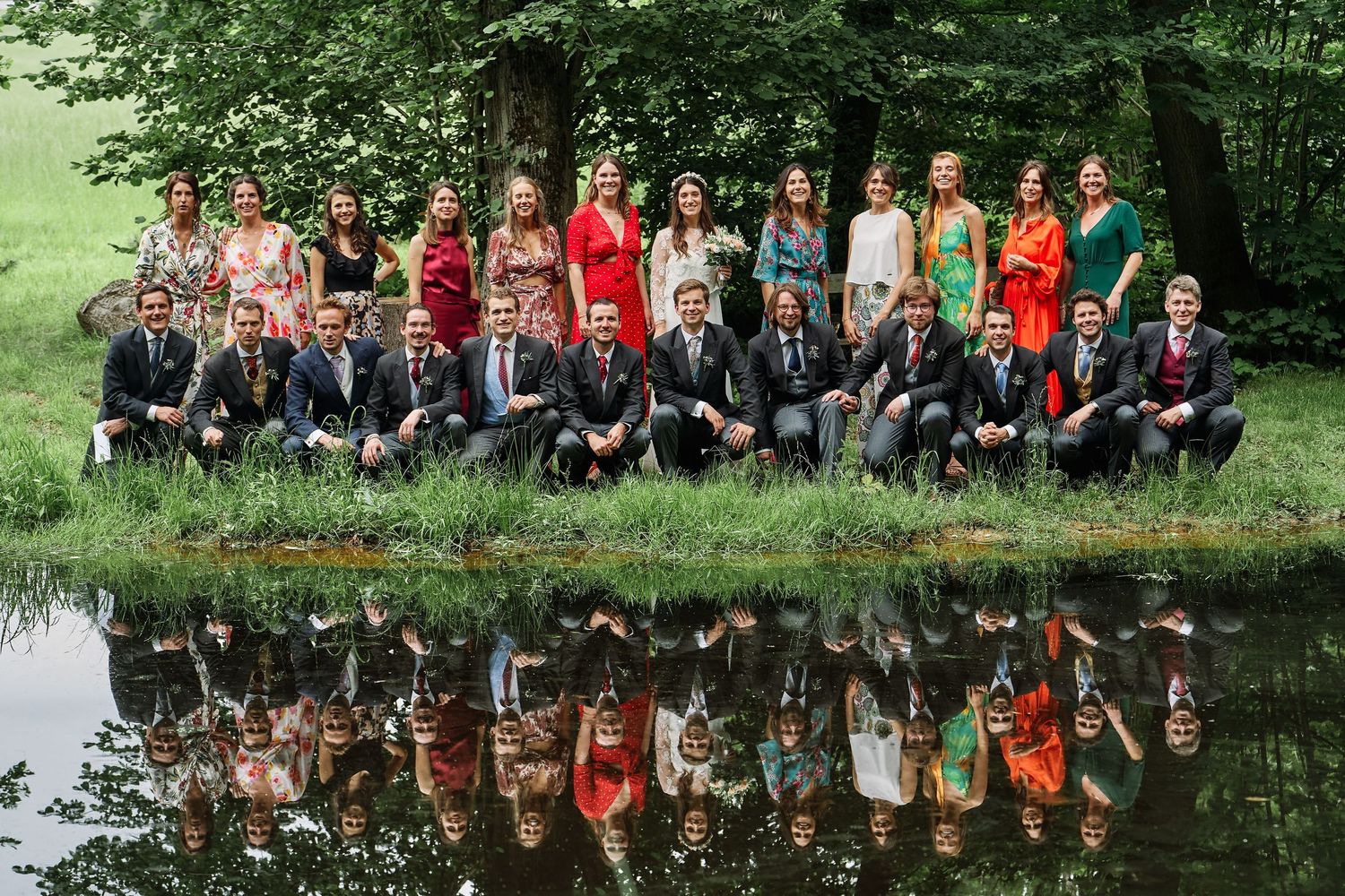 A group of people sitting on a wooden bench outdoors with trees and a reflective pond in the background.