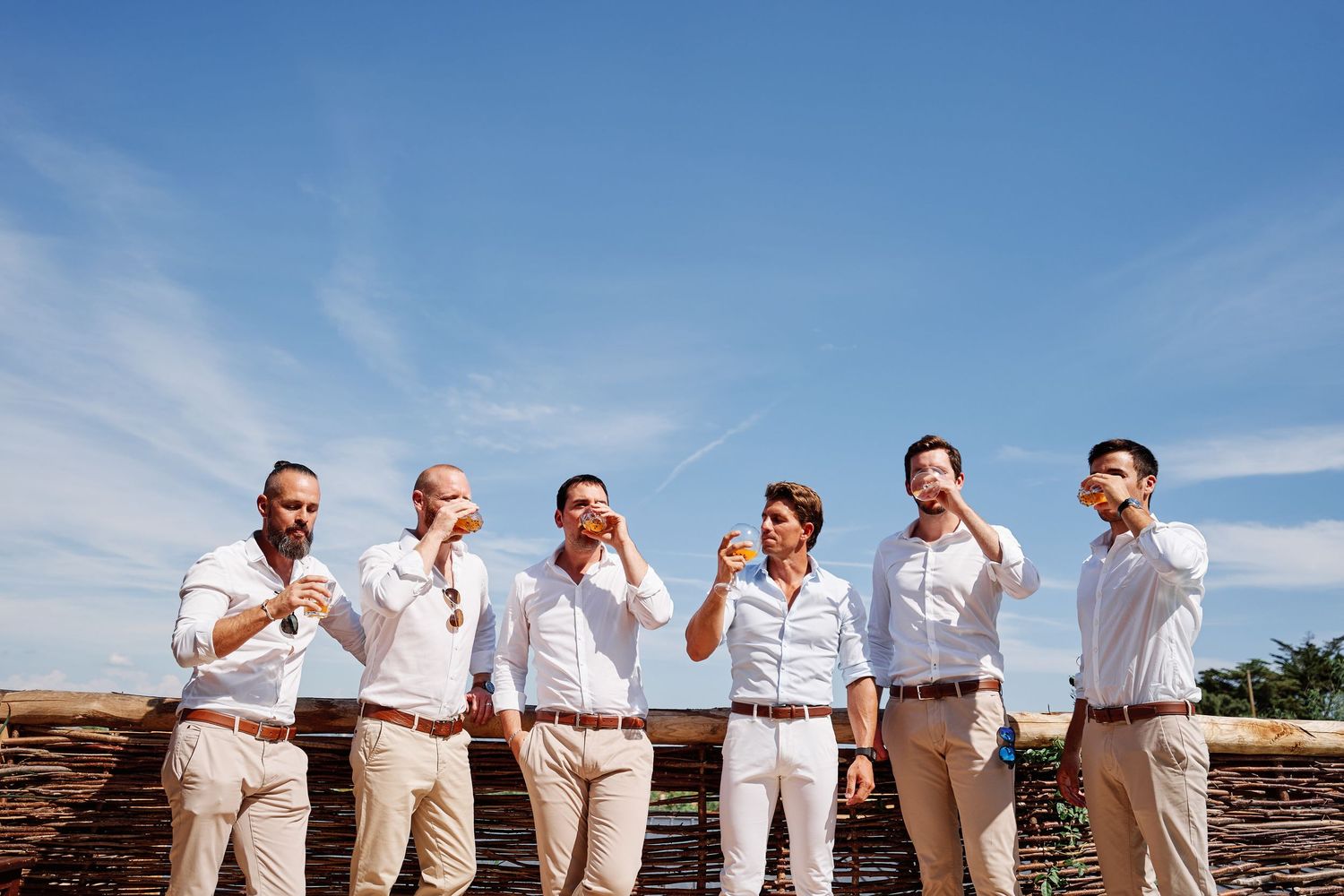 A group of groomsmen in light colored attire pose playfully while covering their faces against a blue sky.