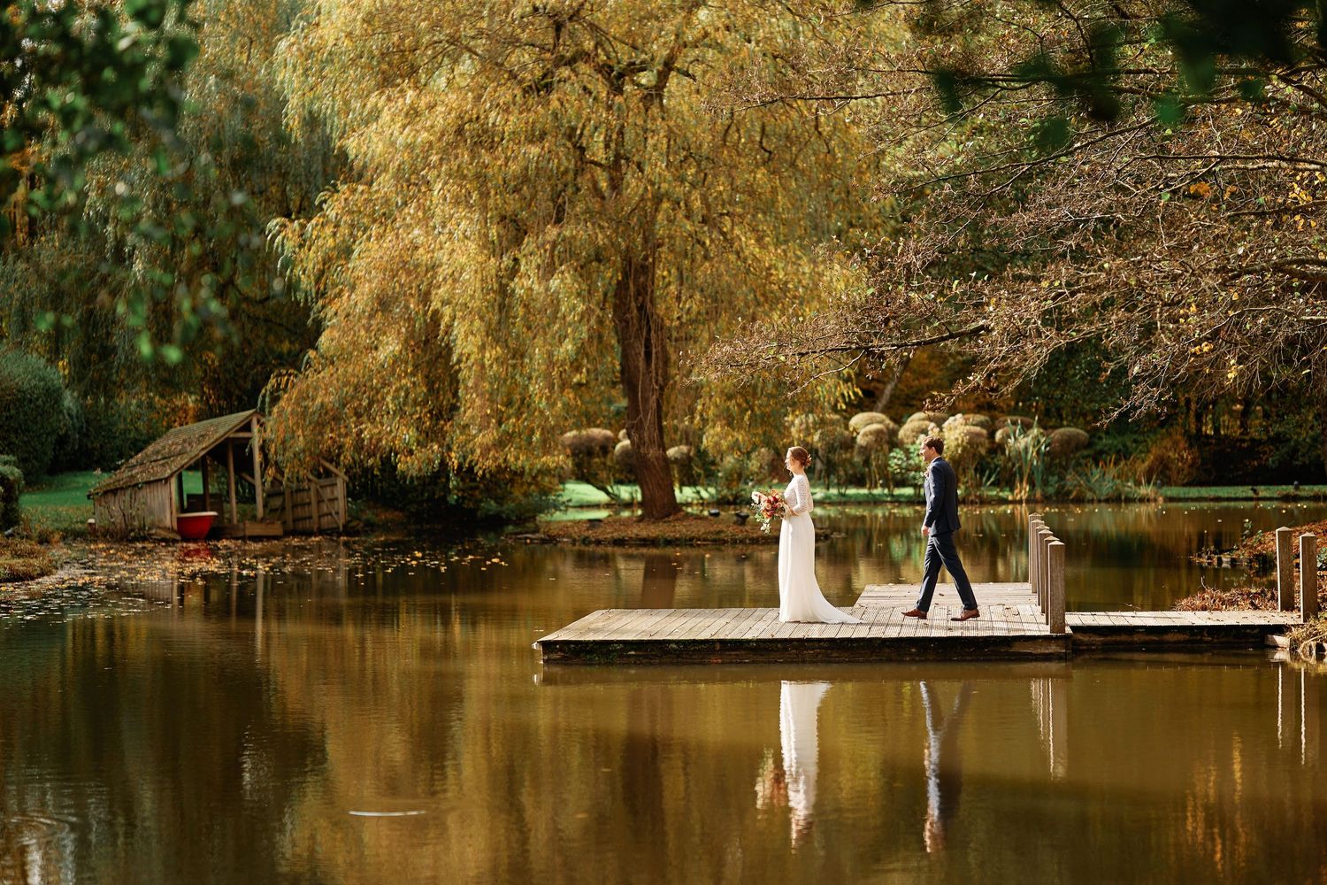 Golden autumn trees reflect in a tranquil lake with wooden docks and a white sailboat in the distance.