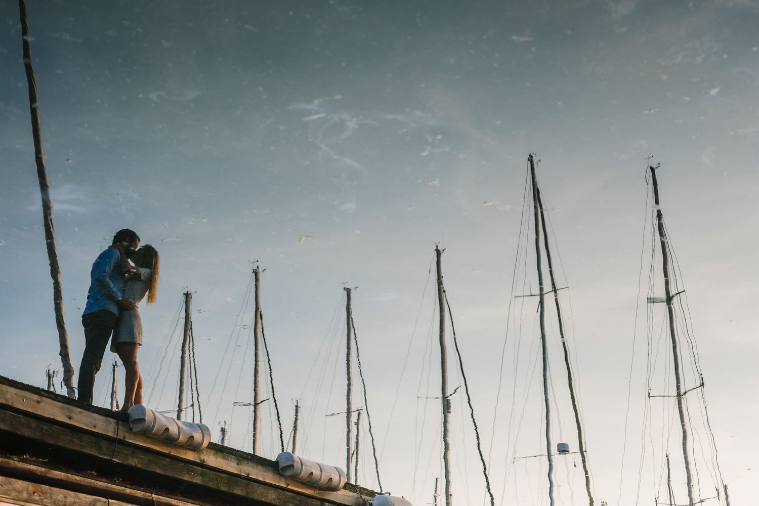 A silhouetted couple stands on a pier with sailboat masts reflecting in the water at sunset.
