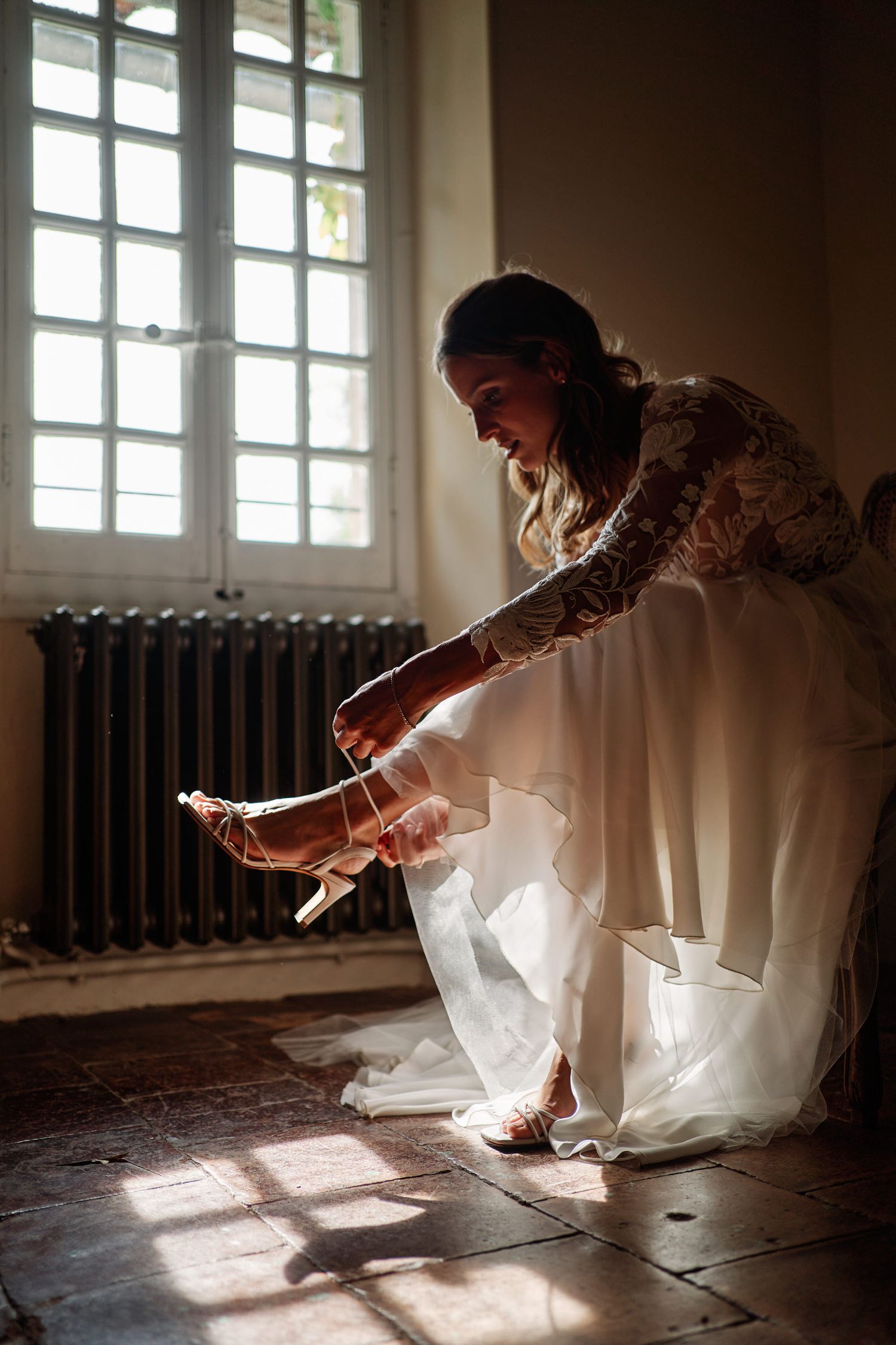 A bride in a white dress crouches down to examine something on the floor in a sunlit room with vintage radiators.