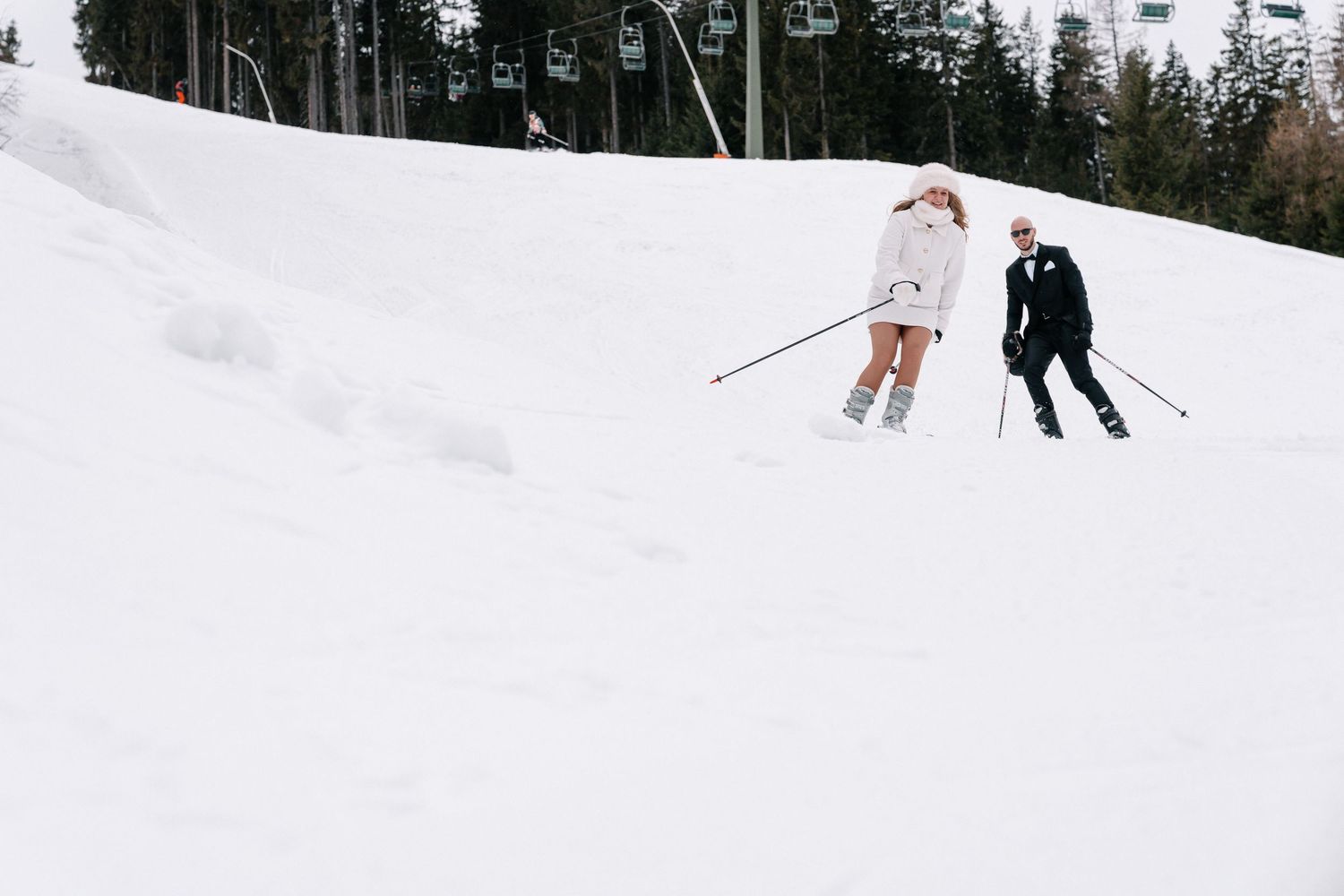 Skiers glide down a snowy mountain slope with evergreen trees and a chairlift visible in the background.