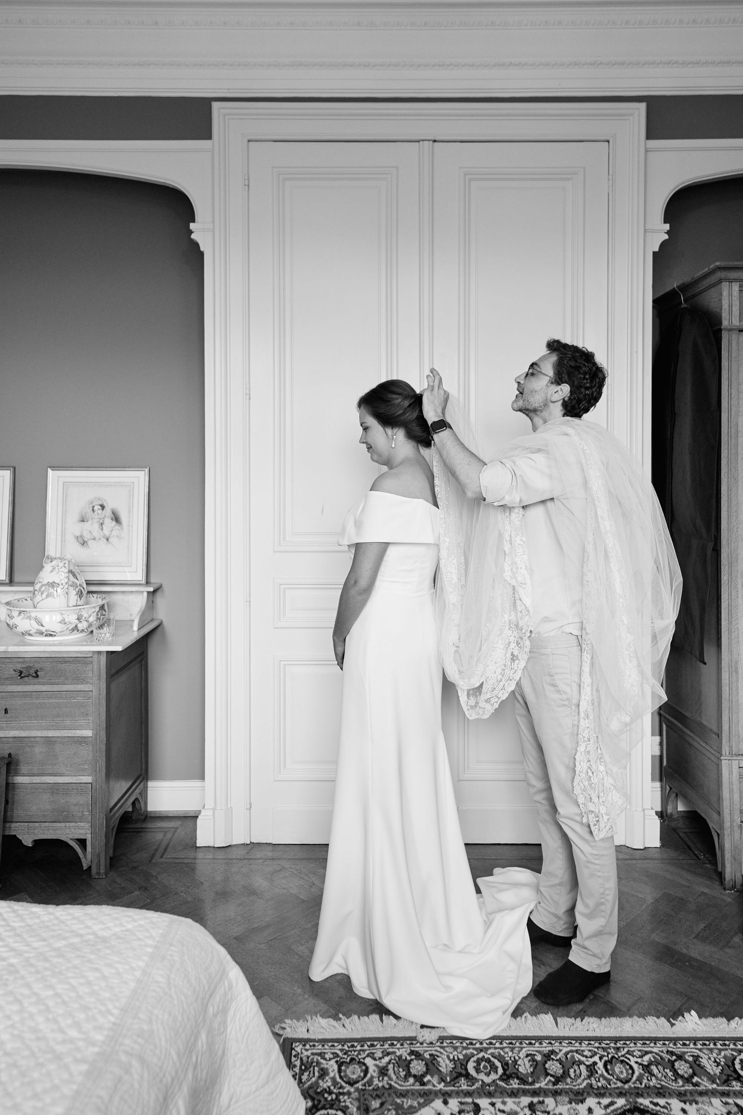A black and white photo capturing a tender moment of wedding preparation in an elegant bedroom setting.
