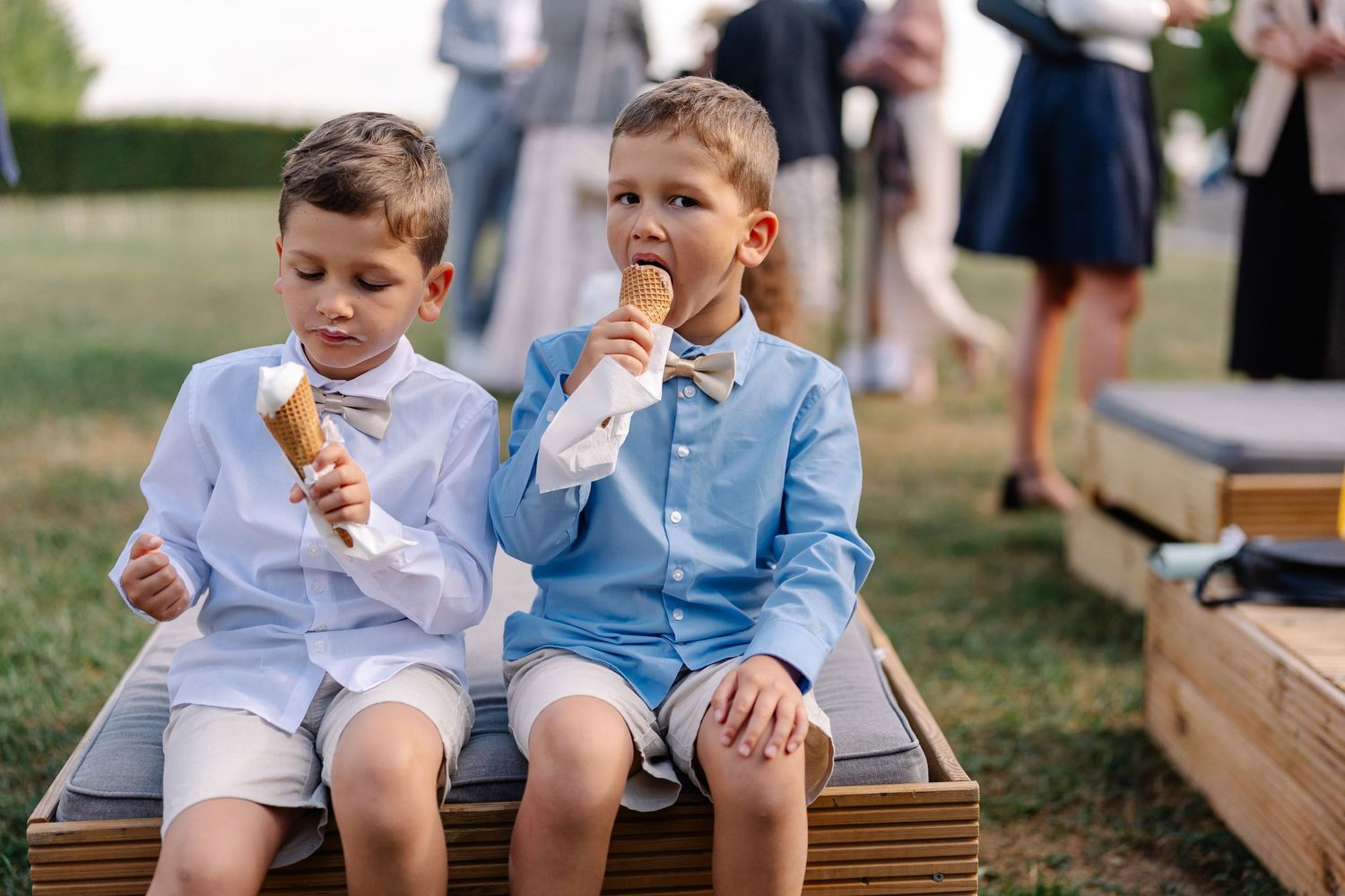 Two children sit together on wooden benches while enjoying ice cream cones on a sunny day.