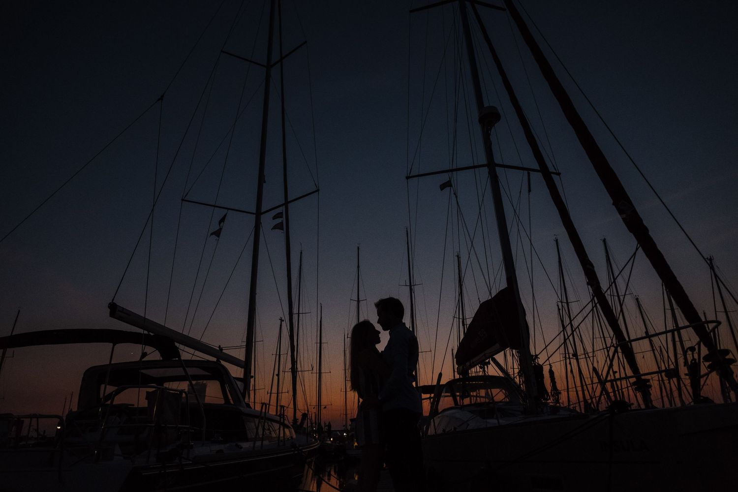 Silhouettes of sailboat masts against a dramatic sunset sky create a striking maritime scene at a harbor.