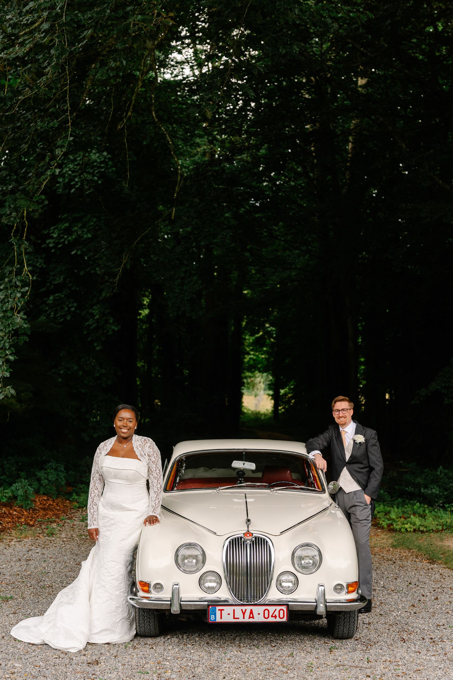 Elegant wedding couple poses with classic white Rolls Royce car on gravel driveway surrounded by lush trees.