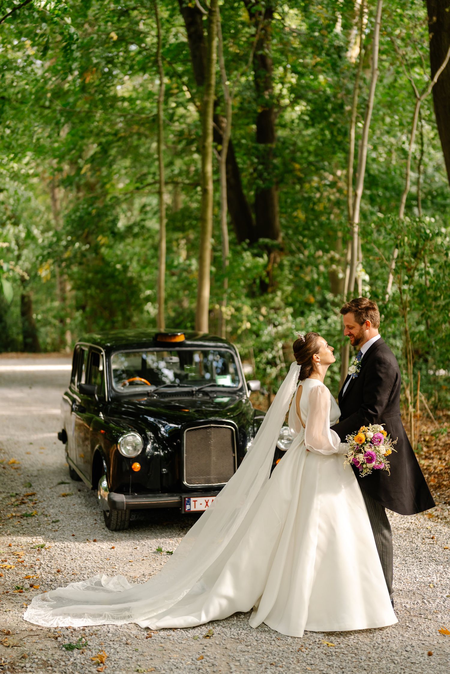 A couple poses near a vintage black car in a wooded setting during their wedding photoshoot.