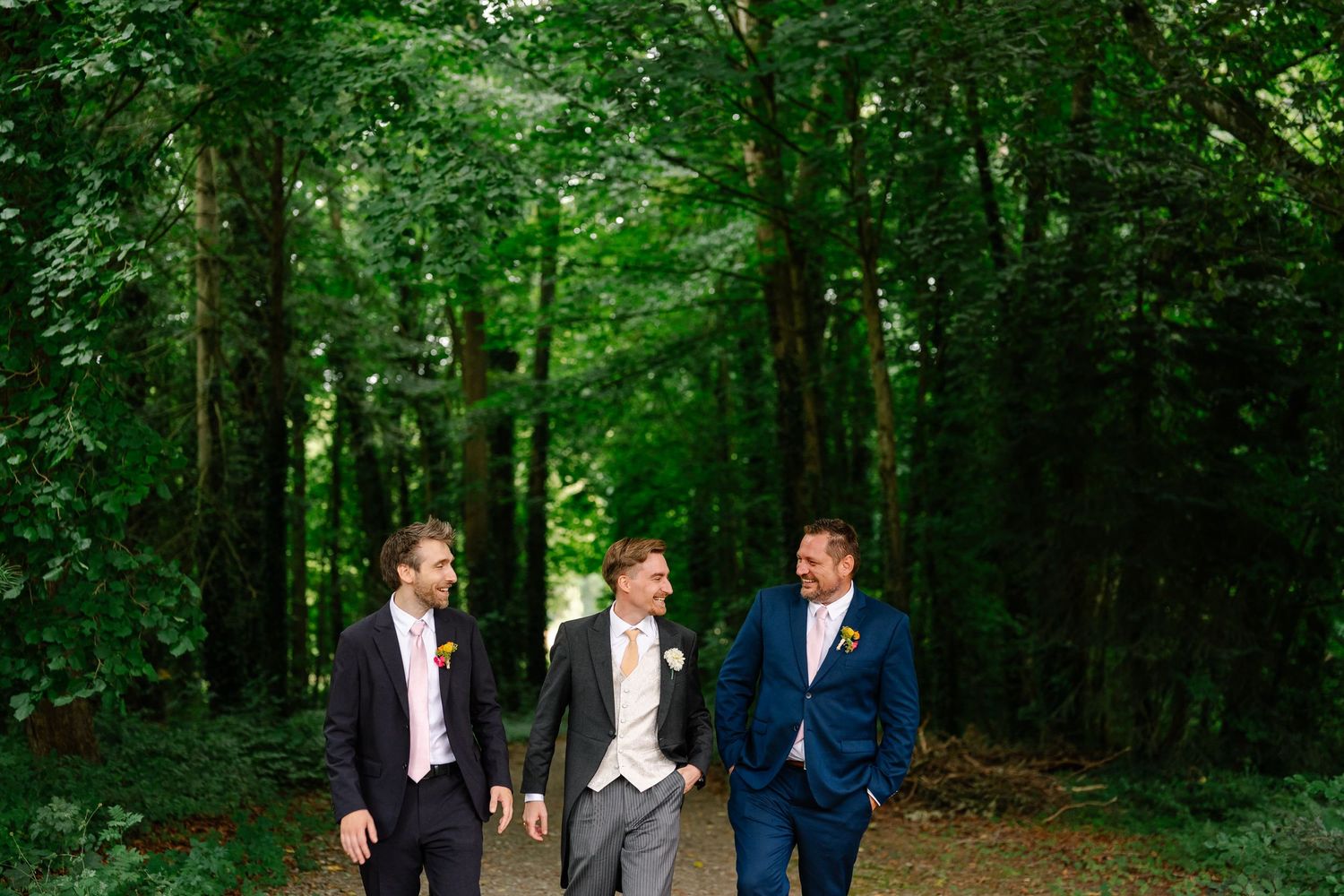 Three groomsmen walk together on a wooded path surrounded by lush green trees during a summer wedding.
