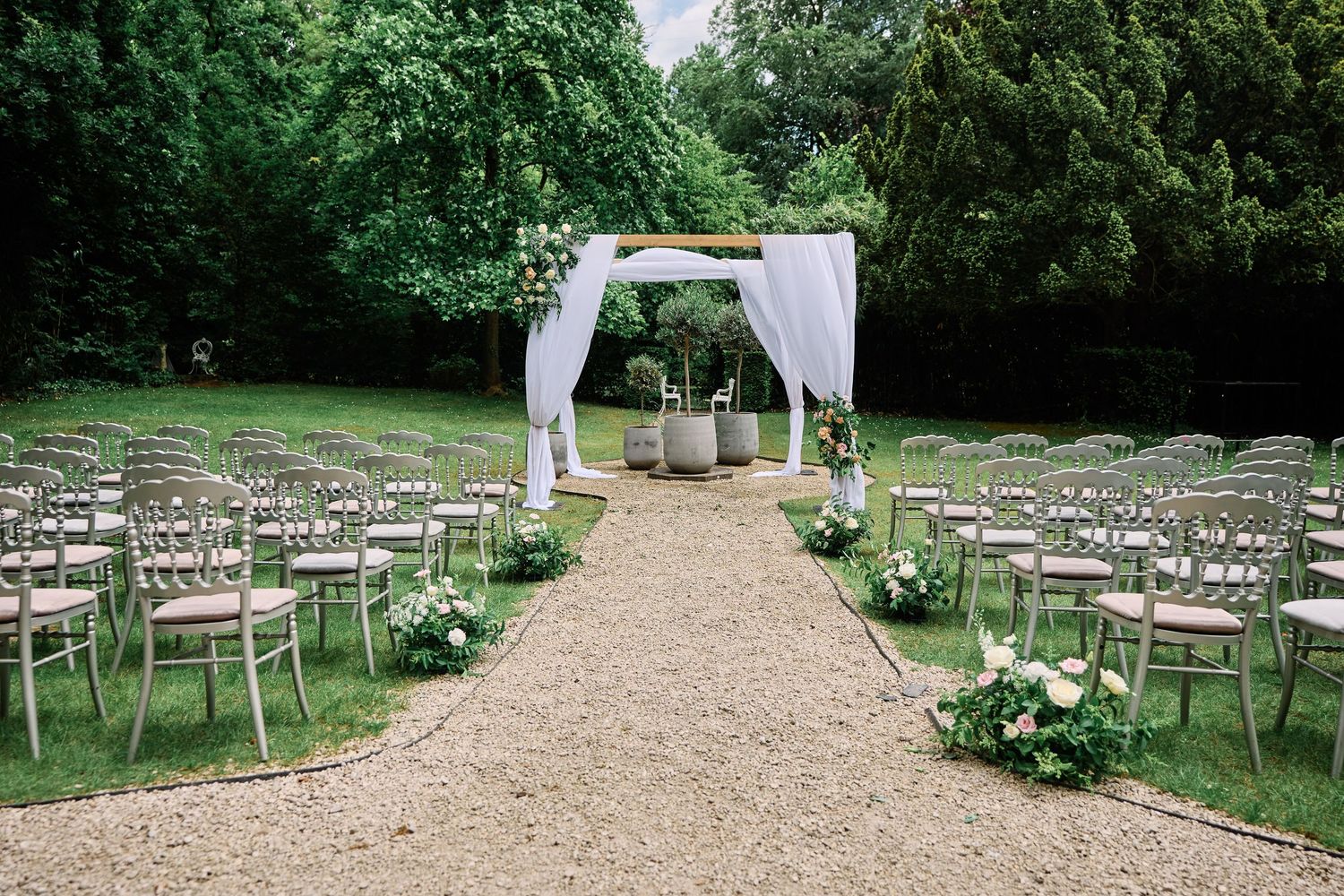 Outdoor wedding ceremony setup with wooden chairs arranged in rows along a gravel path leading to a white arch.