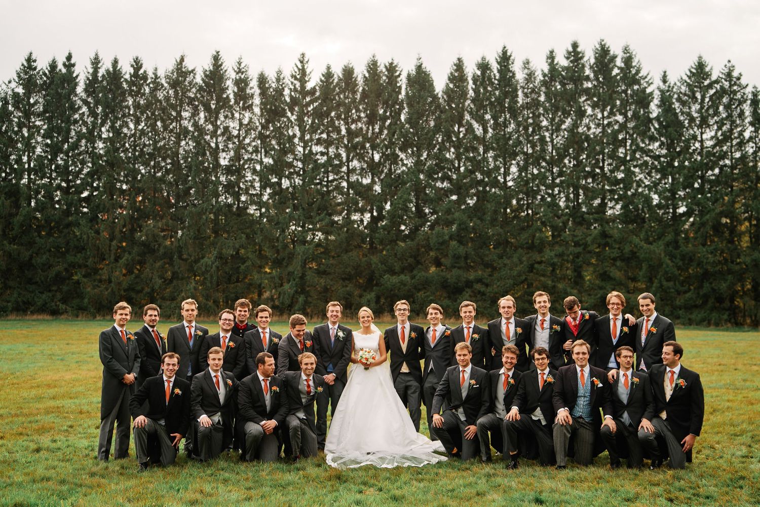 A wedding party in formal attire poses outdoors with tall trees in the background during a sunny day.