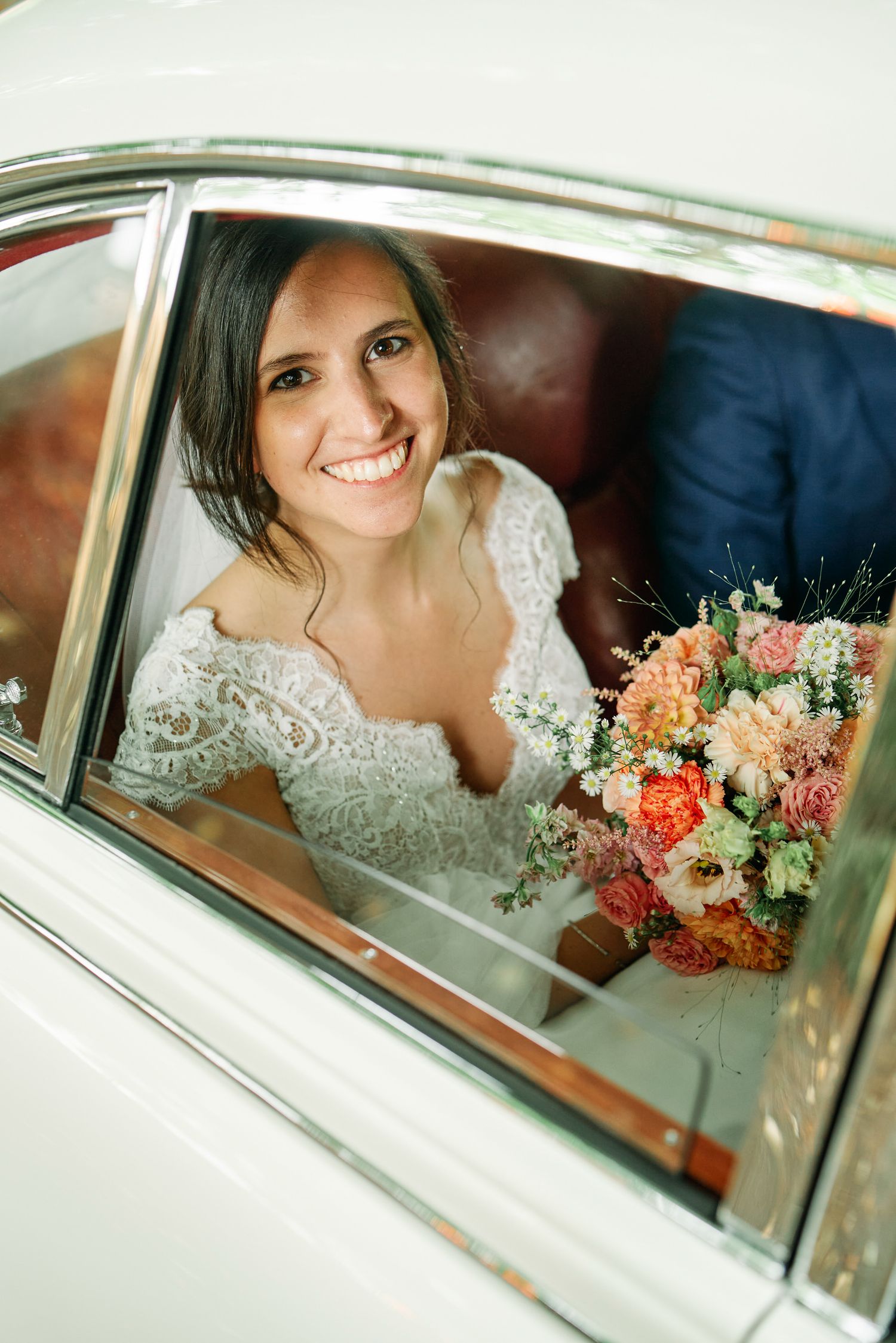 A bride in a lace wedding dress smiles while seated in a vintage car window holding a coral and white floral bouquet.