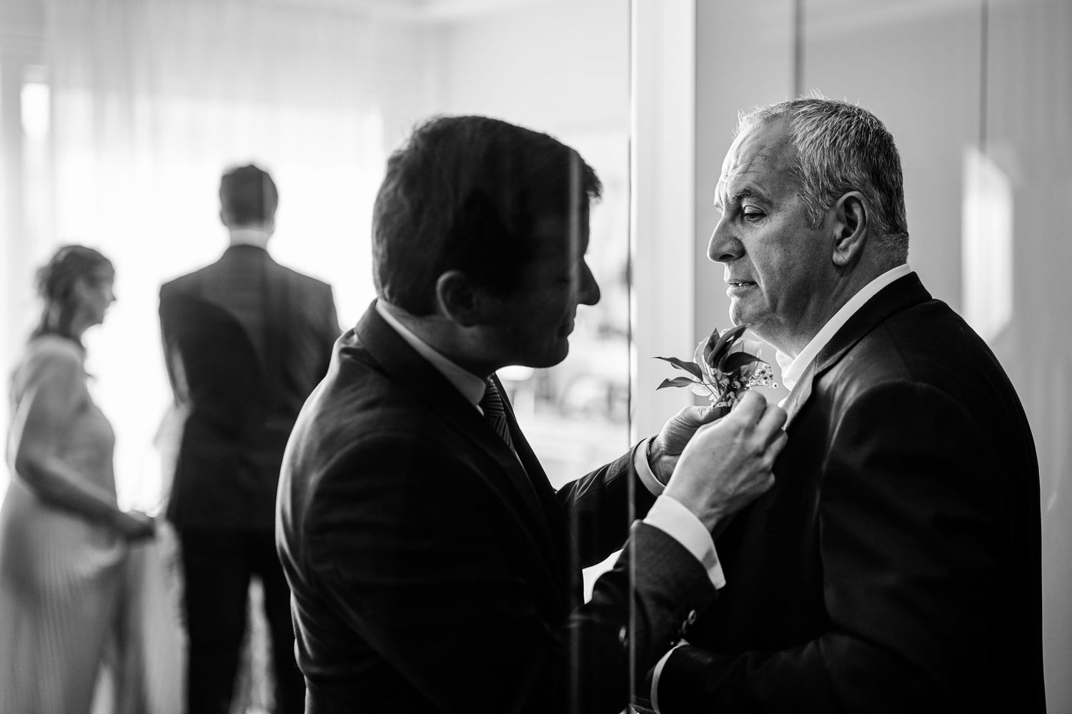 Two men engaged in conversation in a hallway, with silhouettes of other people visible in the background.