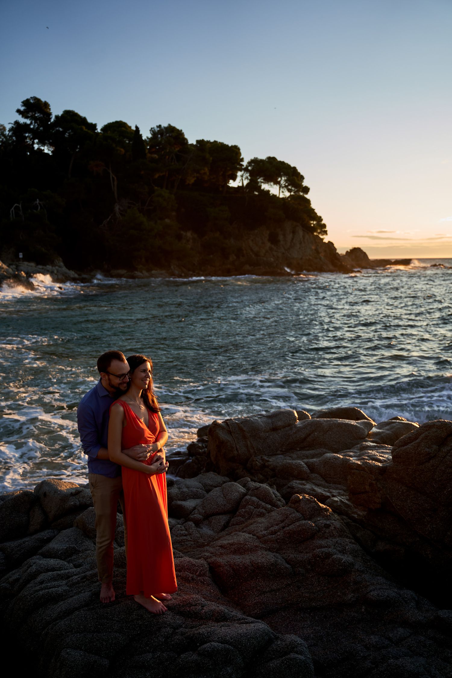 Couple embraces on rocky coastline at sunset as waves crash against the shore.