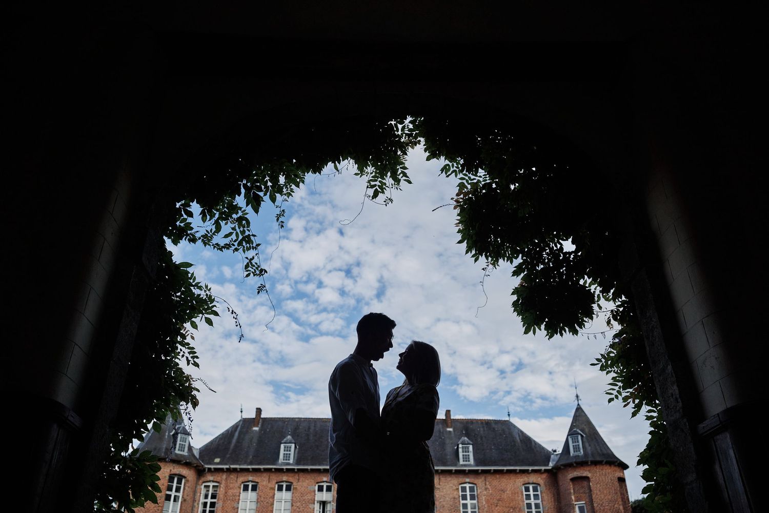 Silhouetted couple framed by trees with historic chateau and cloudy sky in background.
