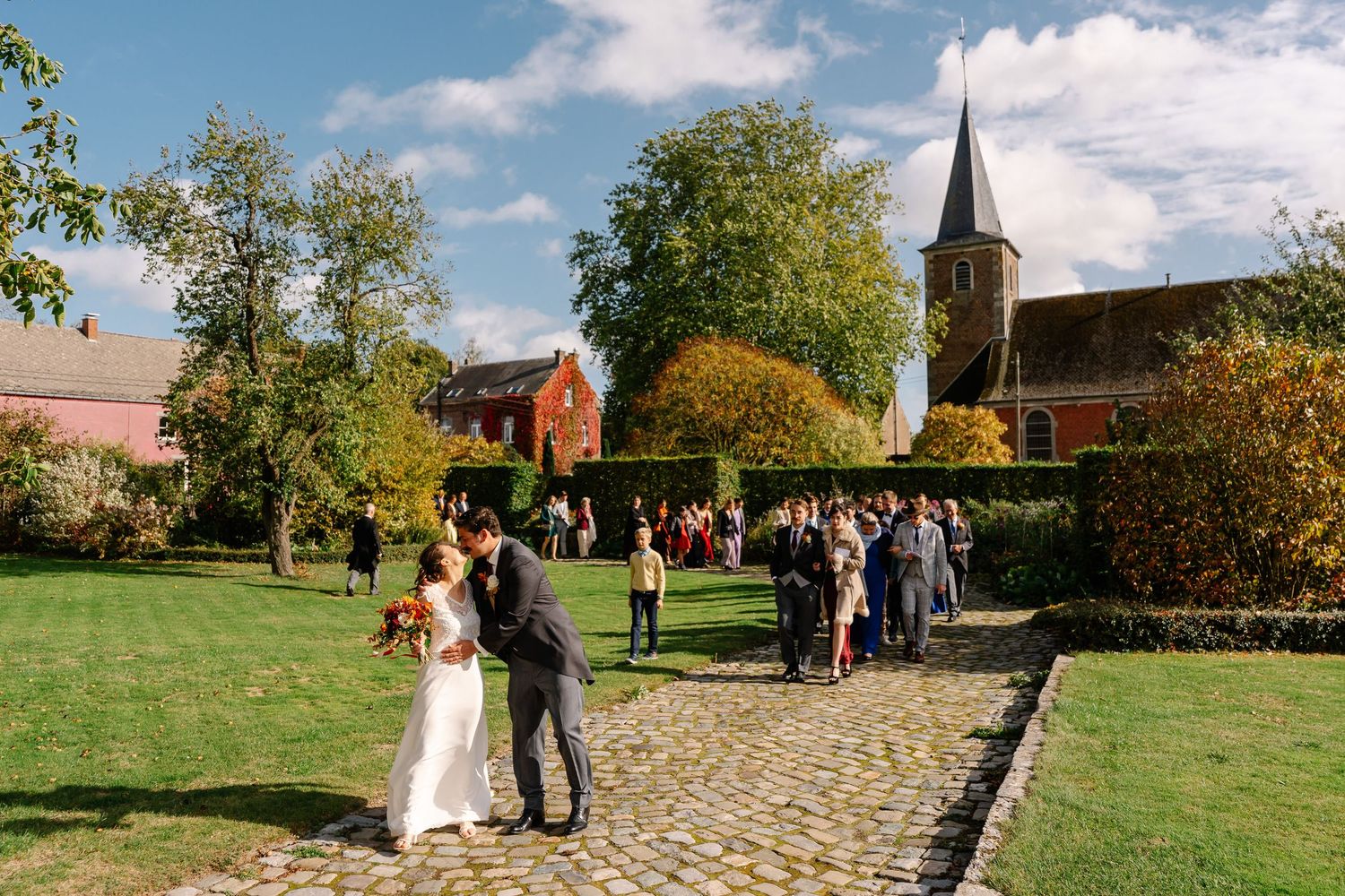 A beautiful outdoor wedding ceremony takes place in a rustic countryside setting with a church steeple in the background.