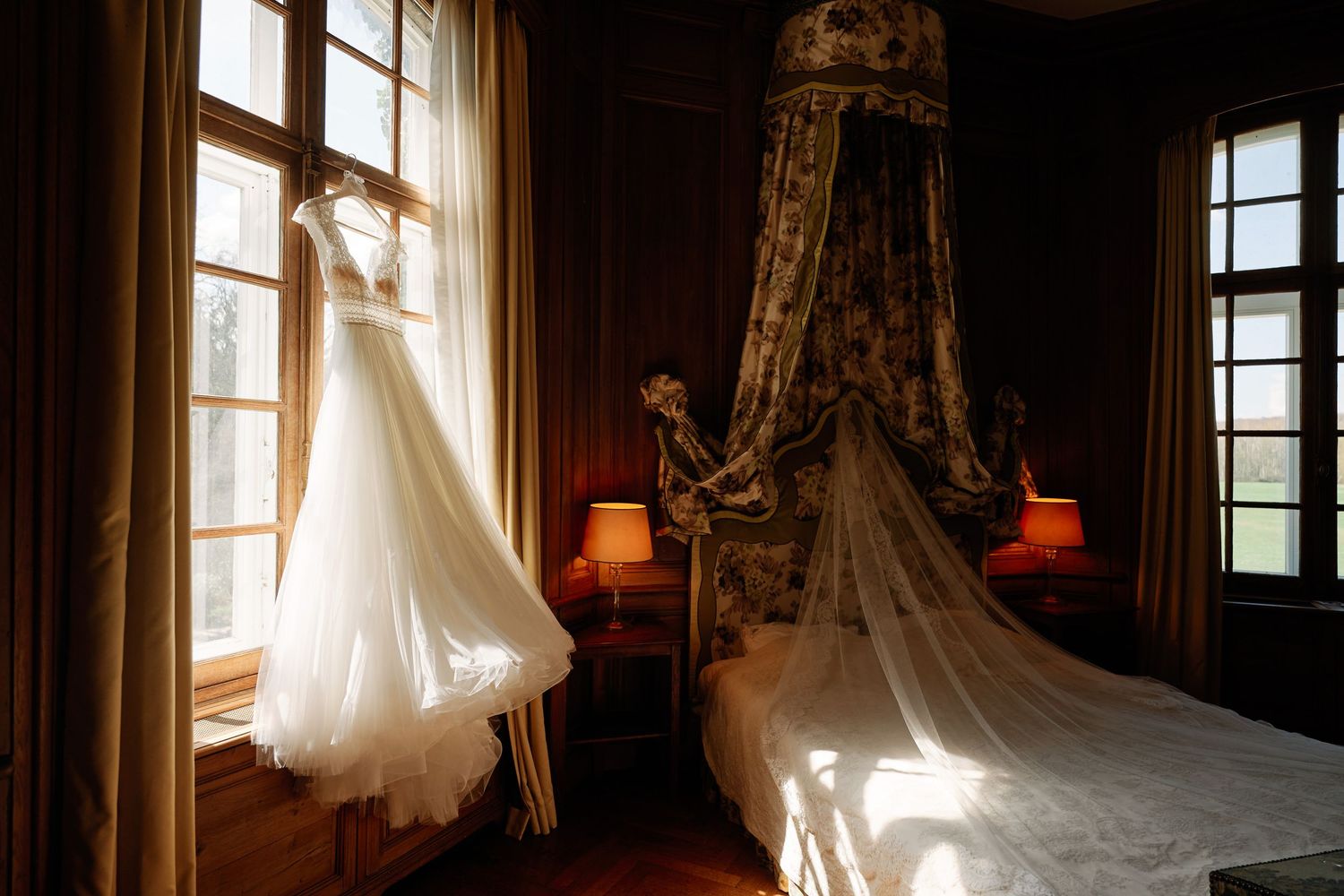 Sunlight streams through windows illuminating a white wedding dress hanging in an elegant room.