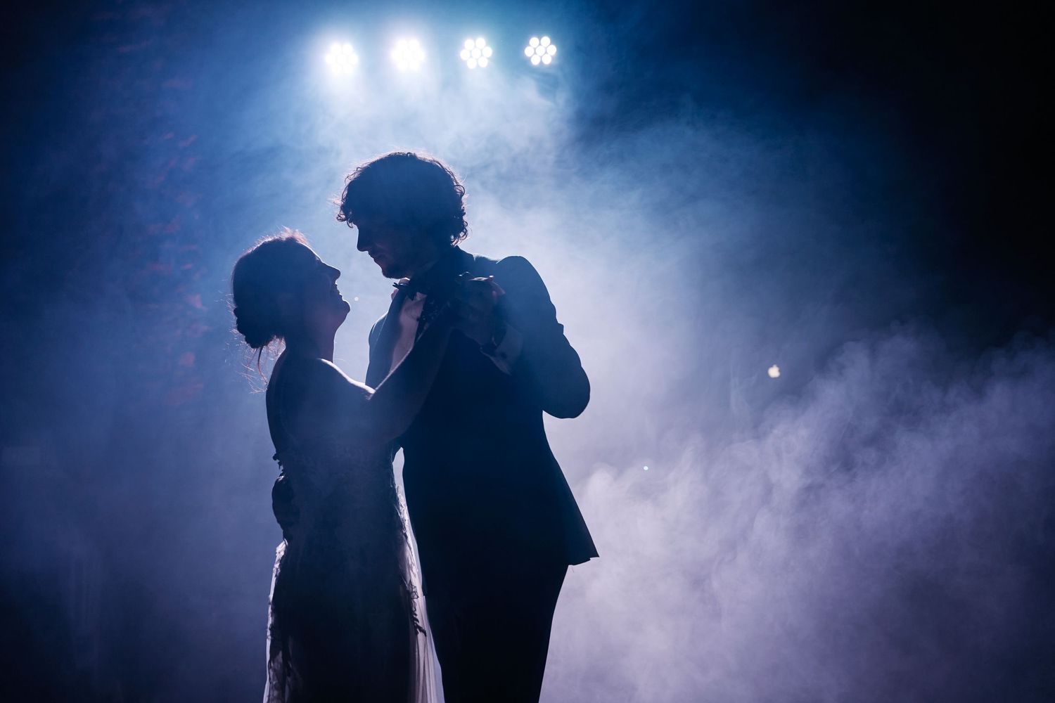Silhouetted figures perform on a smoky stage with bright white spotlights creating a dramatic backlit effect.