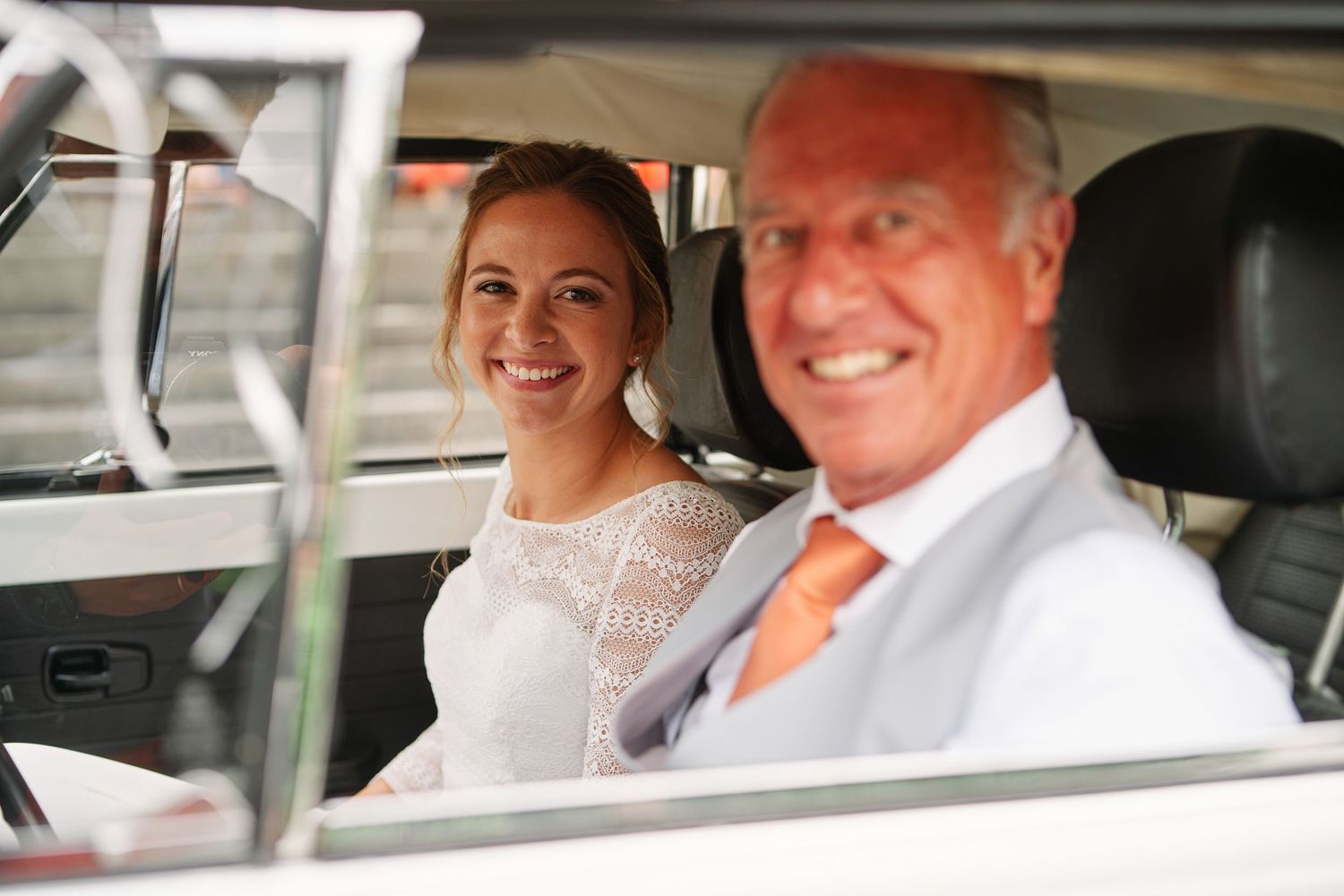 Two people sit in a car with white interior while smiling at the camera through the windshield.
