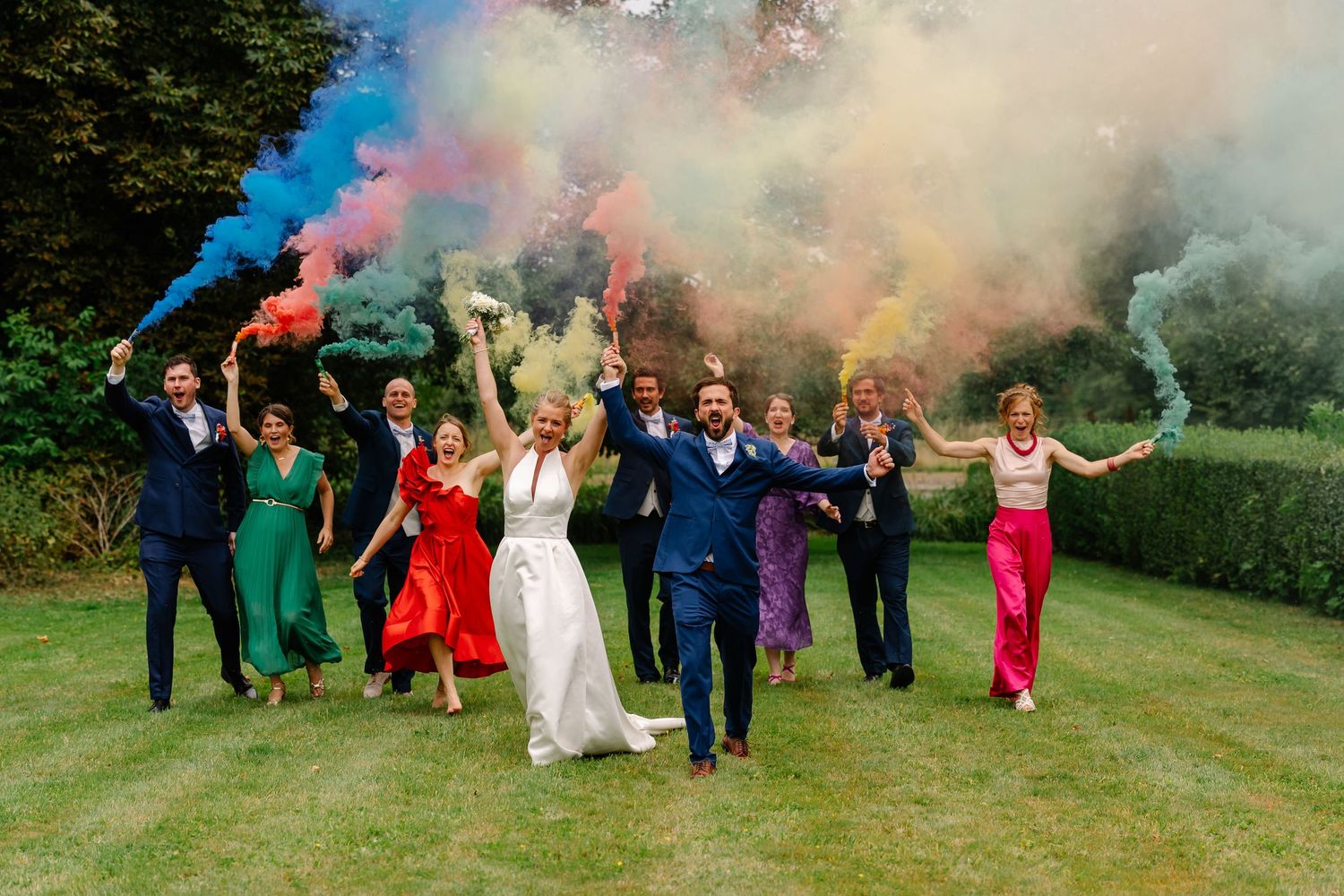 Wedding party celebrates with colorful smoke bombs in an outdoor garden setting creating vibrant photo effects.