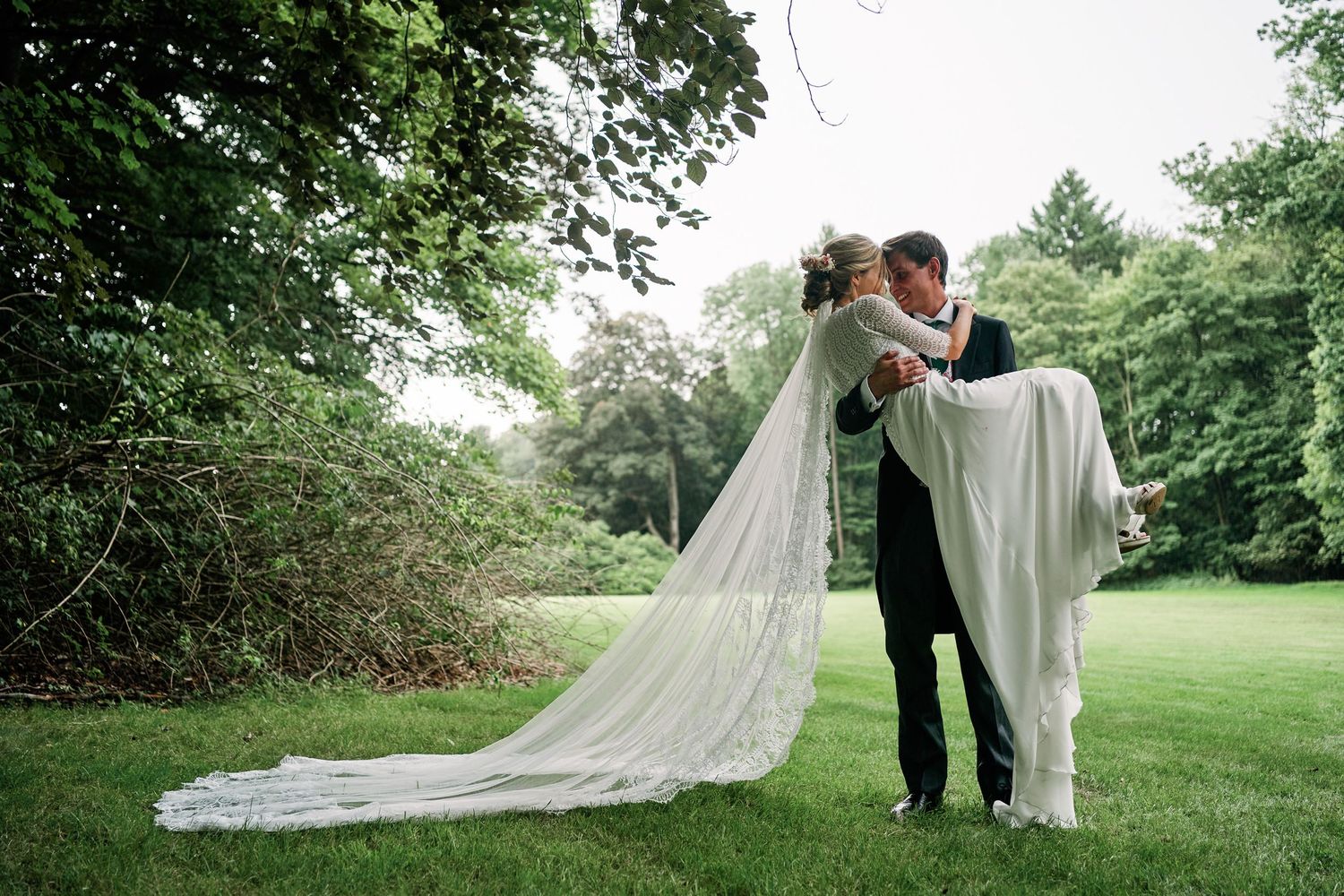 A romantic wedding couple embraces on a lush green lawn with a flowing long veil trailing behind them.