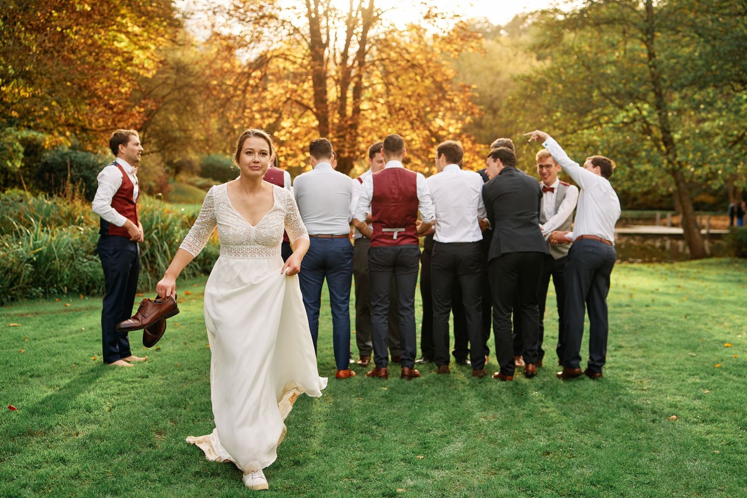 Wedding party gathers outdoors in a park at sunset during golden hour for group photos.