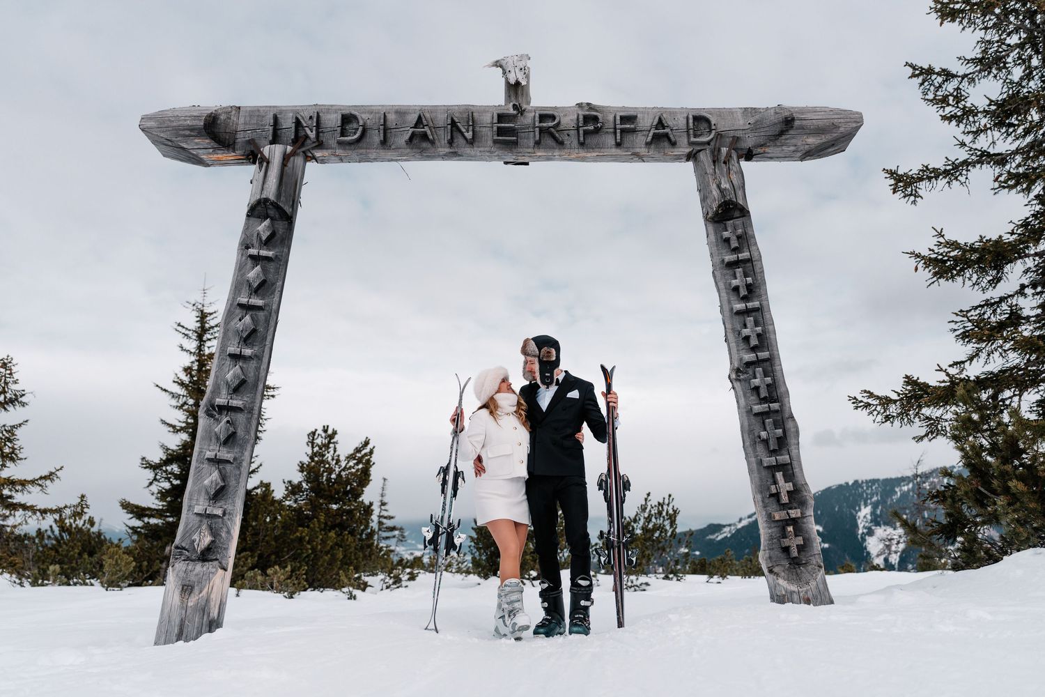 Winter wedding photo series with couple posing under wooden Indian Head sign in snowy mountain landscape.
