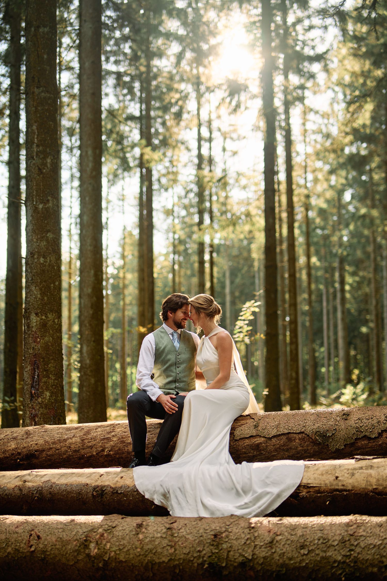 Newlywed couple sits romantically on fallen log in sunlit forest during golden hour wedding photoshoot.
