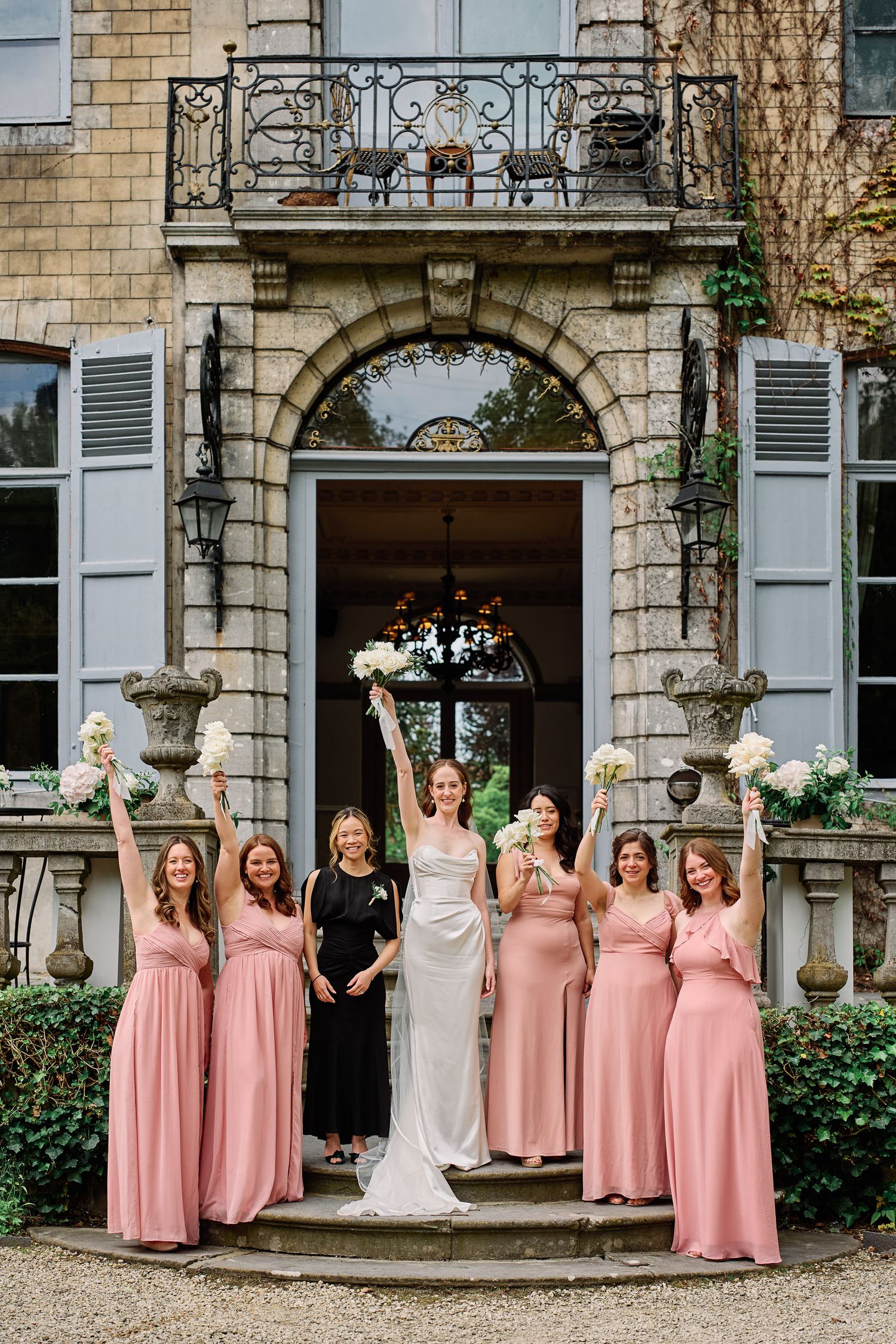 A wedding party in pink bridesmaid dresses poses together outside an elegant stone building with white shutters.