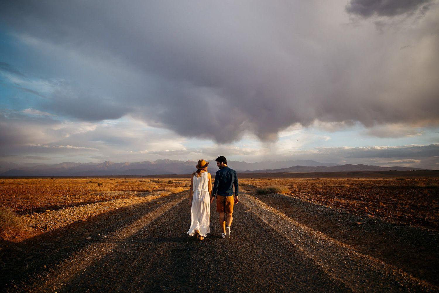 Couple walking along a dirt road through an arid desert landscape under dramatic stormy clouds at sunset.