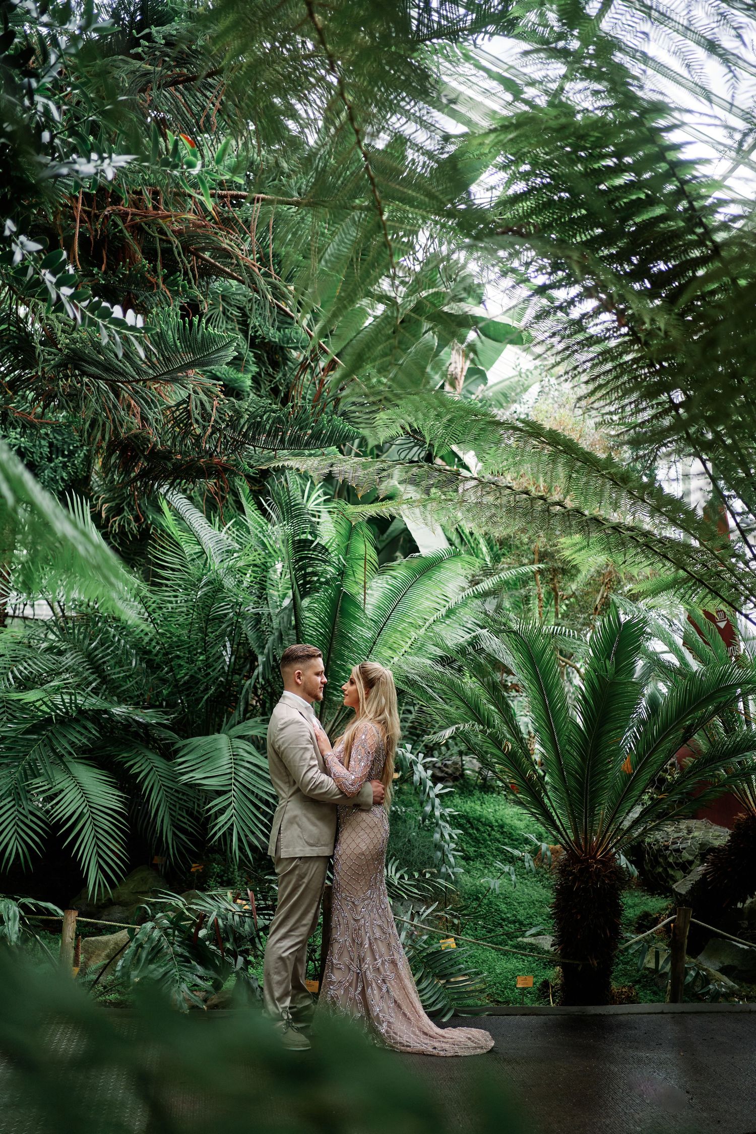 A romantic couple embraces in an elegant greenhouse setting surrounded by lush tropical ferns and palm fronds.