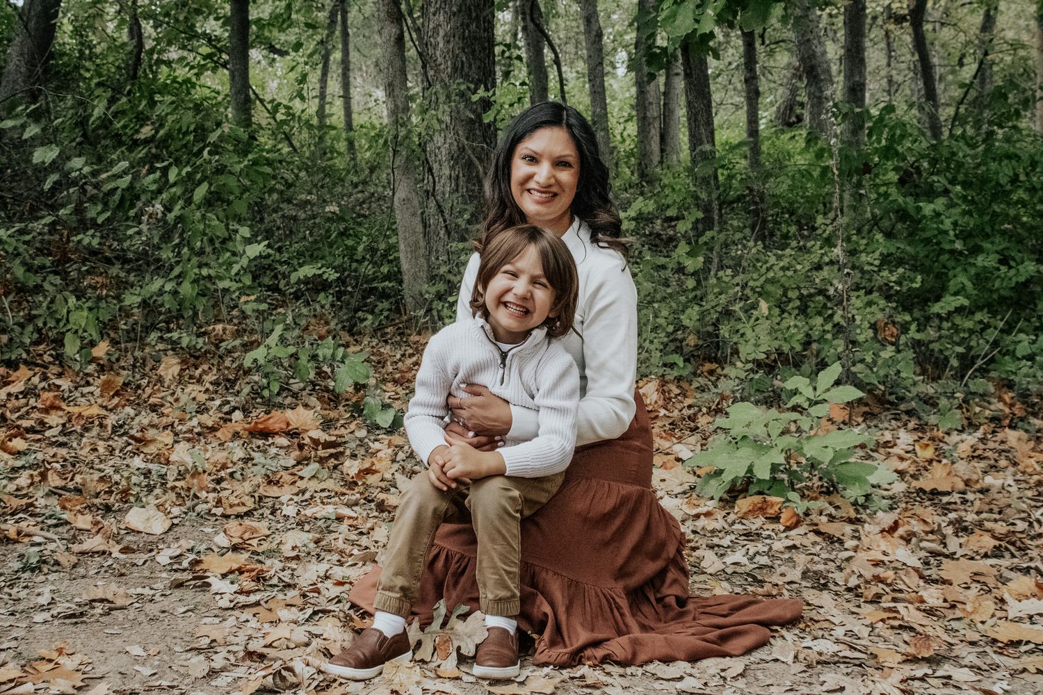 Smiling mother sitting on autumn leaves with her young son in a wooded outdoor setting.