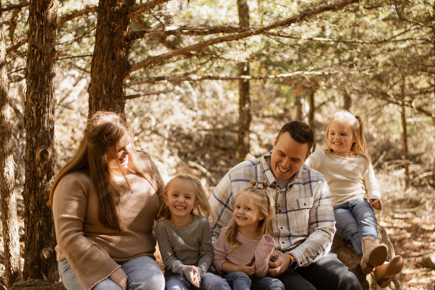 Happy family of five sitting together and laughing among pine trees in a beautiful forest setting.