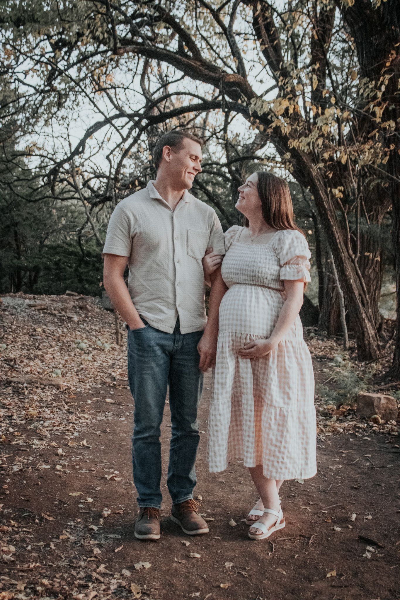 Smiling pregnant couple standing together under trees on a leaf-covered path outdoors.