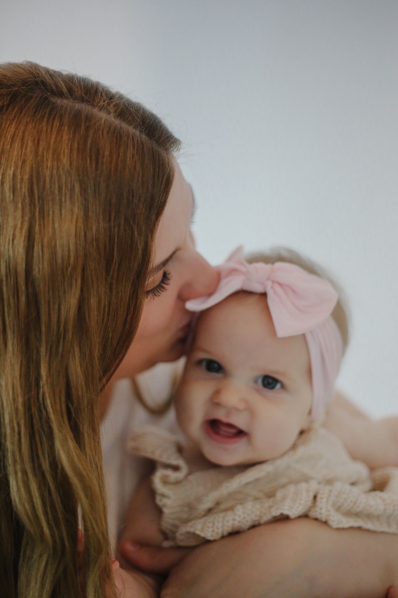 A mother tenderly kisses her smiling baby girl wearing a pink bow headband.