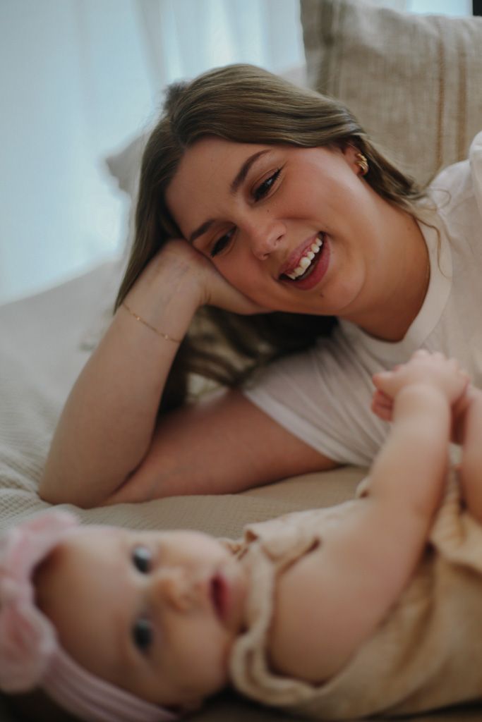 A smiling young mother lying on a couch gazing lovingly at her baby girl wearing a pink headband.