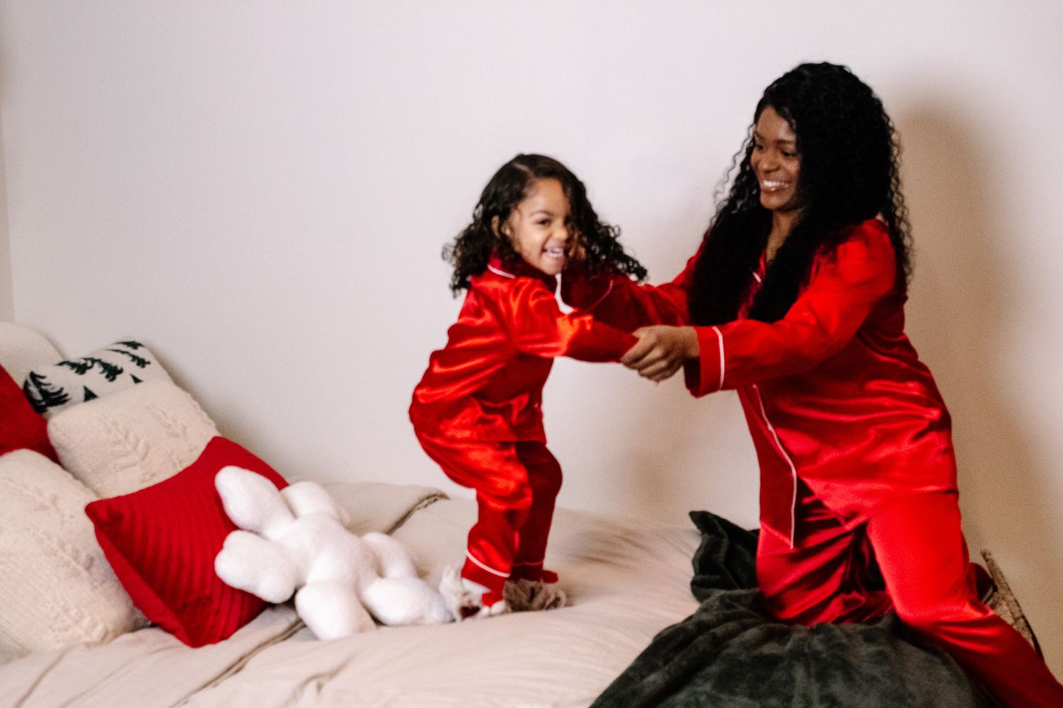 Mother and young daughter dancing on a bed wearing matching red satin Christmas pajamas in a festive holiday photo.