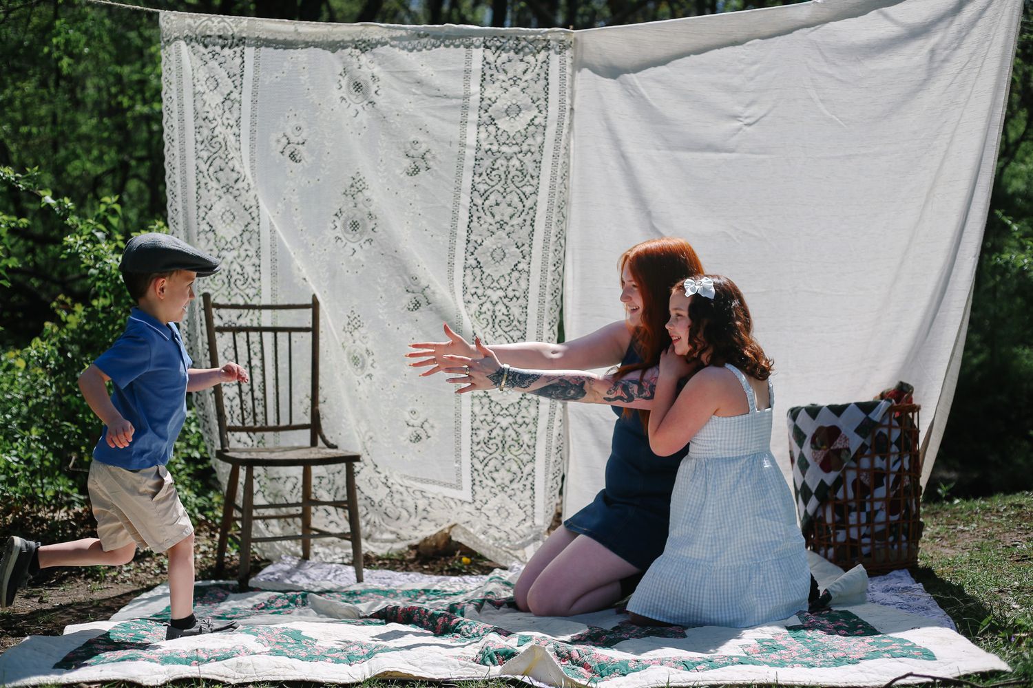 Woman in white dress kneels on quilt outdoors, arms open to welcome running boy near vintage chair and lace backdrop.