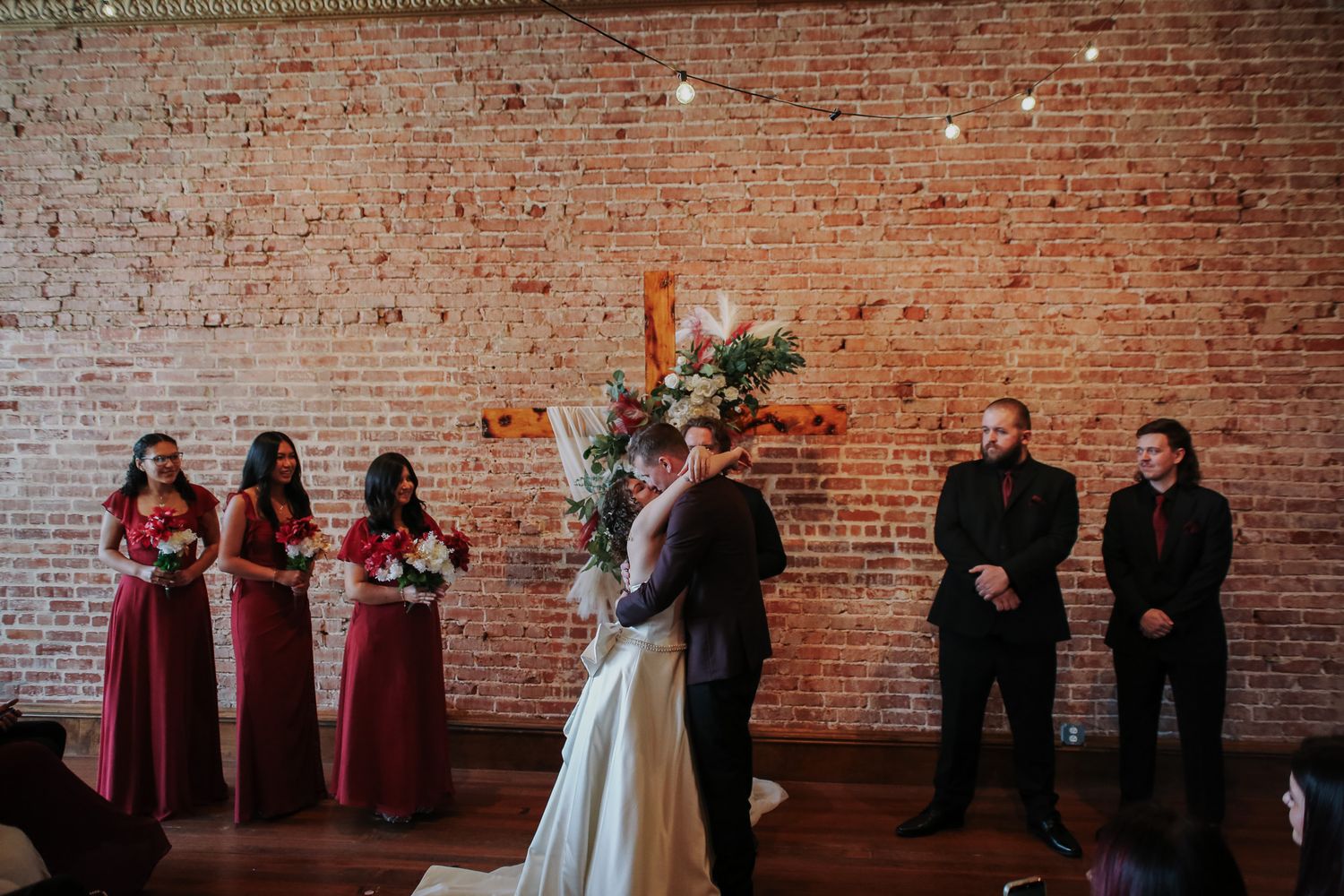 Wedding ceremony first kiss at rustic brick wall venue with bridesmaids in burgundy dresses.